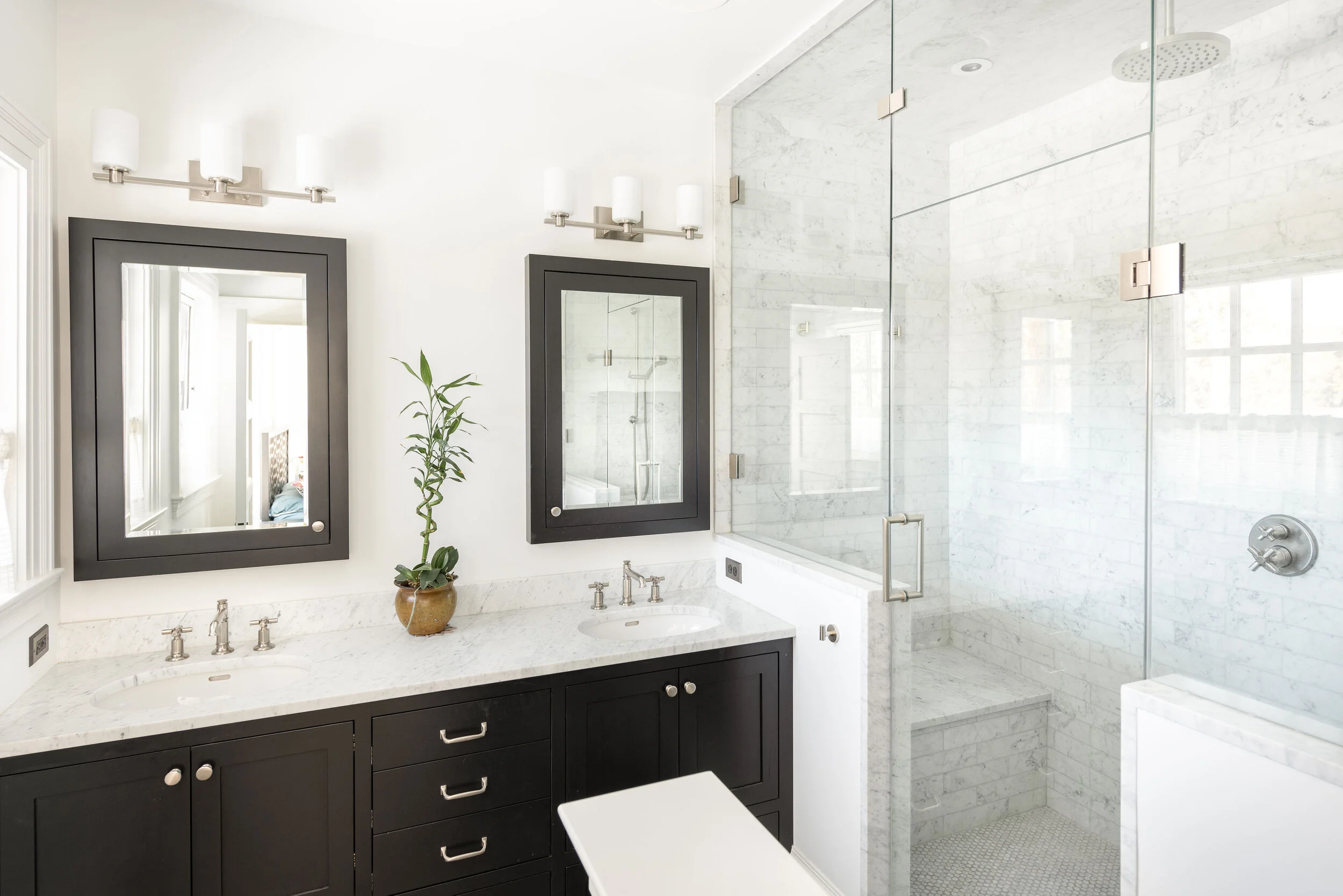 Modern bathroom with dual black-framed mirrors, a potted plant, a marble double vanity, and a glass-enclosed shower with marble walls.