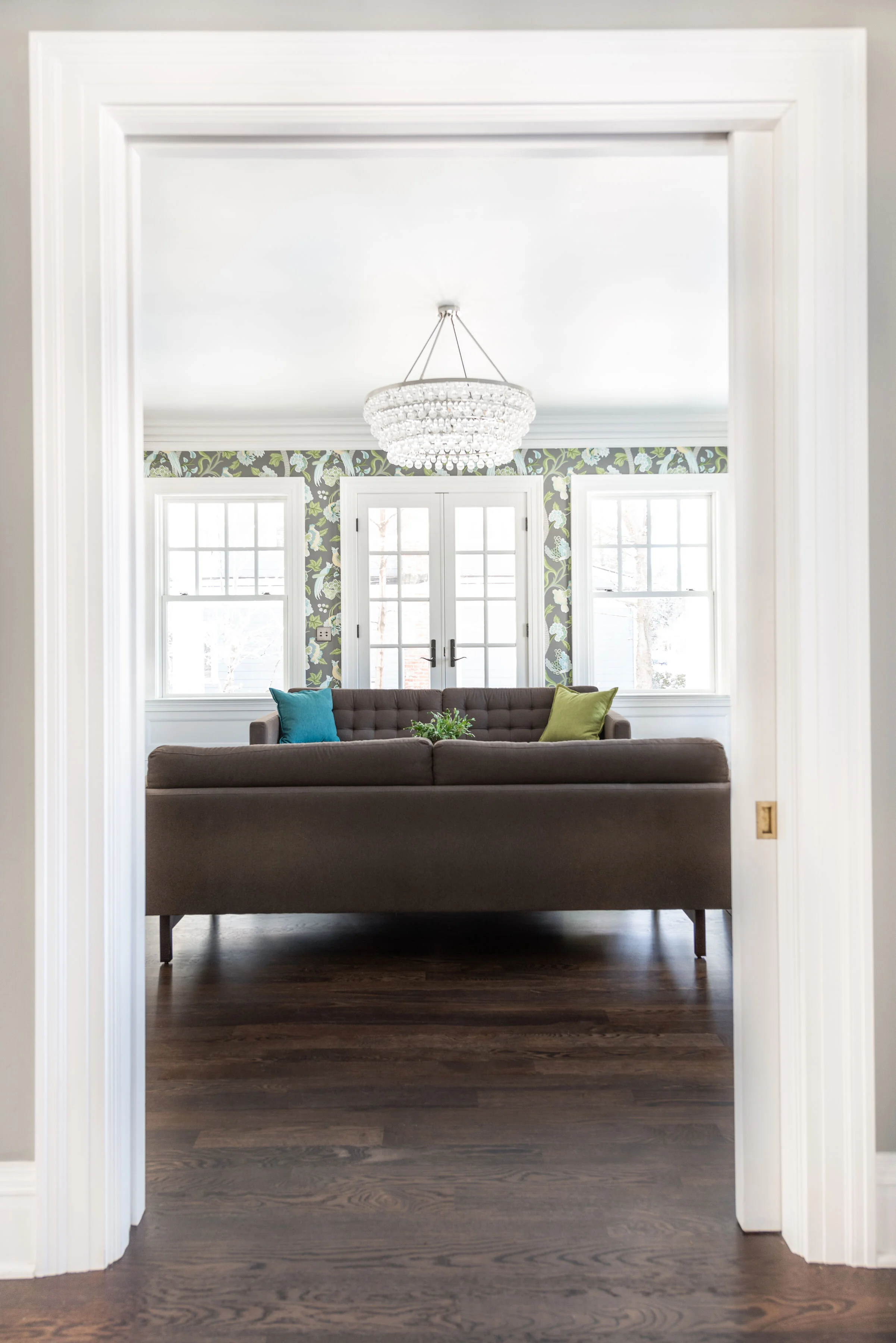 View of a living room seen through a doorway, featuring a dark brown sofa with colorful cushions, a chandelier, two windows, and glass doors with a floral wallpaper background.