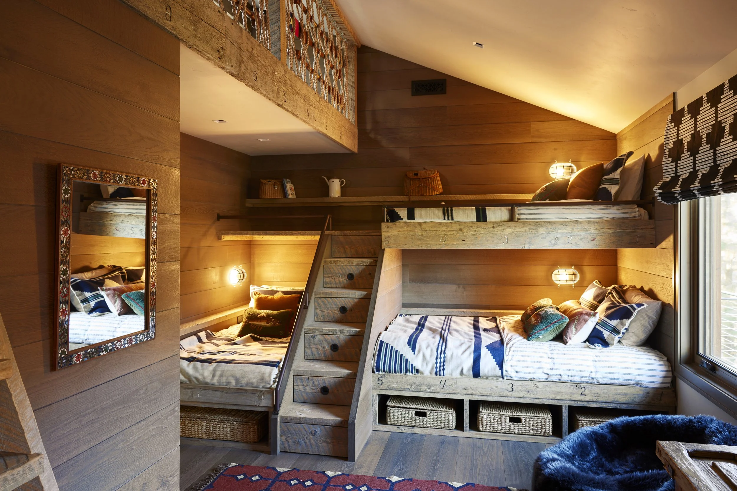 Cozy bedroom with wooden walls featuring bunk beds, a small ladder, and a window with black and white patterned curtains; warm lighting, decorative pillows, woven storage baskets, and a colorful rug.