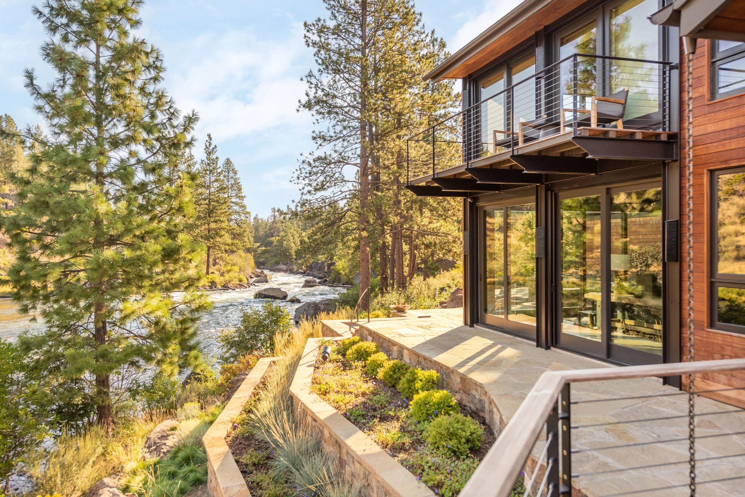 Modern house with large glass sliding doors and a balcony overlooking a river and forested landscape during daytime.