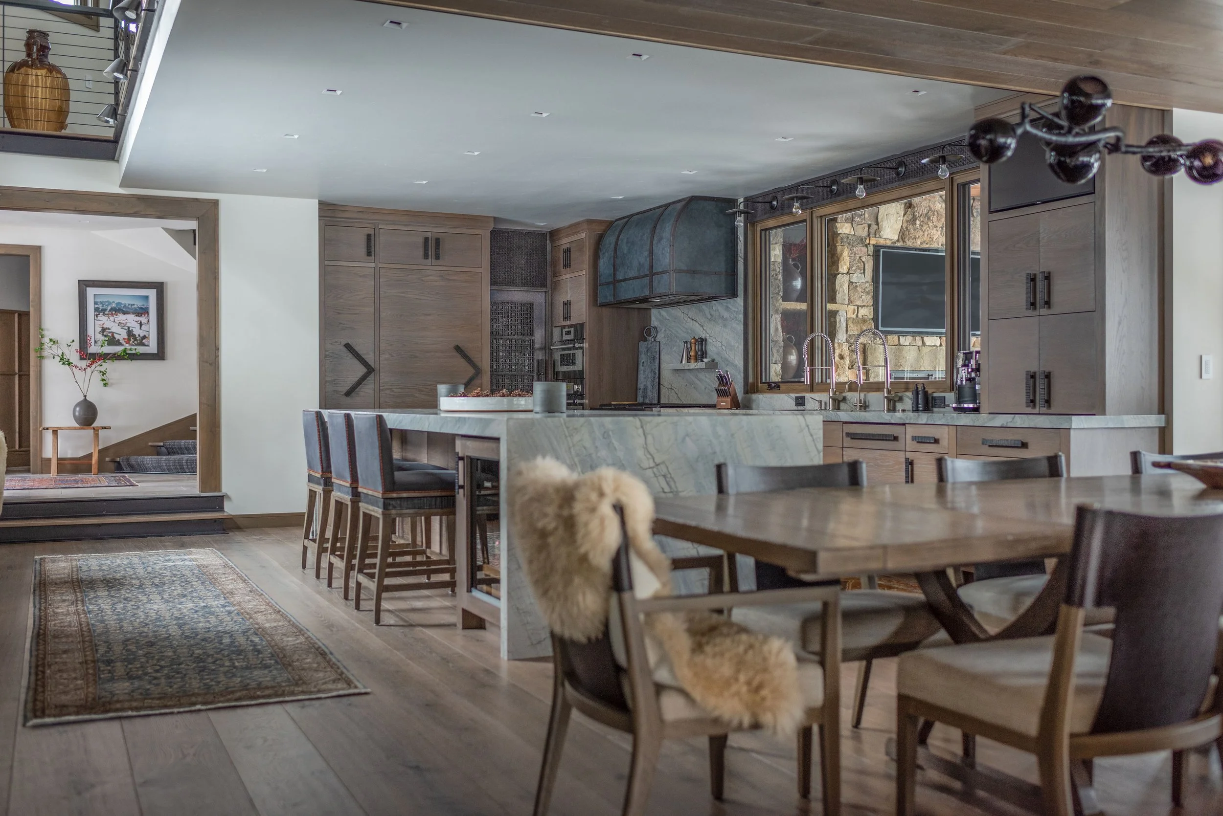 Modern kitchen and dining area with wooden furniture, marble countertops, and large windows showing stone exterior. The dining table has various chairs, one with a sheepskin throw. The kitchen features built-in cabinets, a range hood, and a televisio