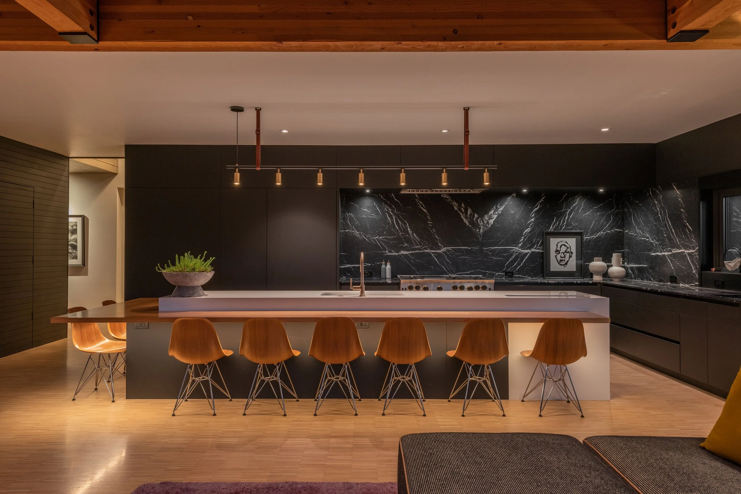 Modern luxury kitchen featuring black cabinets, a dramatic black marble backsplash, exposed timber ceiling beams, and a large island with wood seating in a custom home by Copperline Homes in Bend, Oregon.