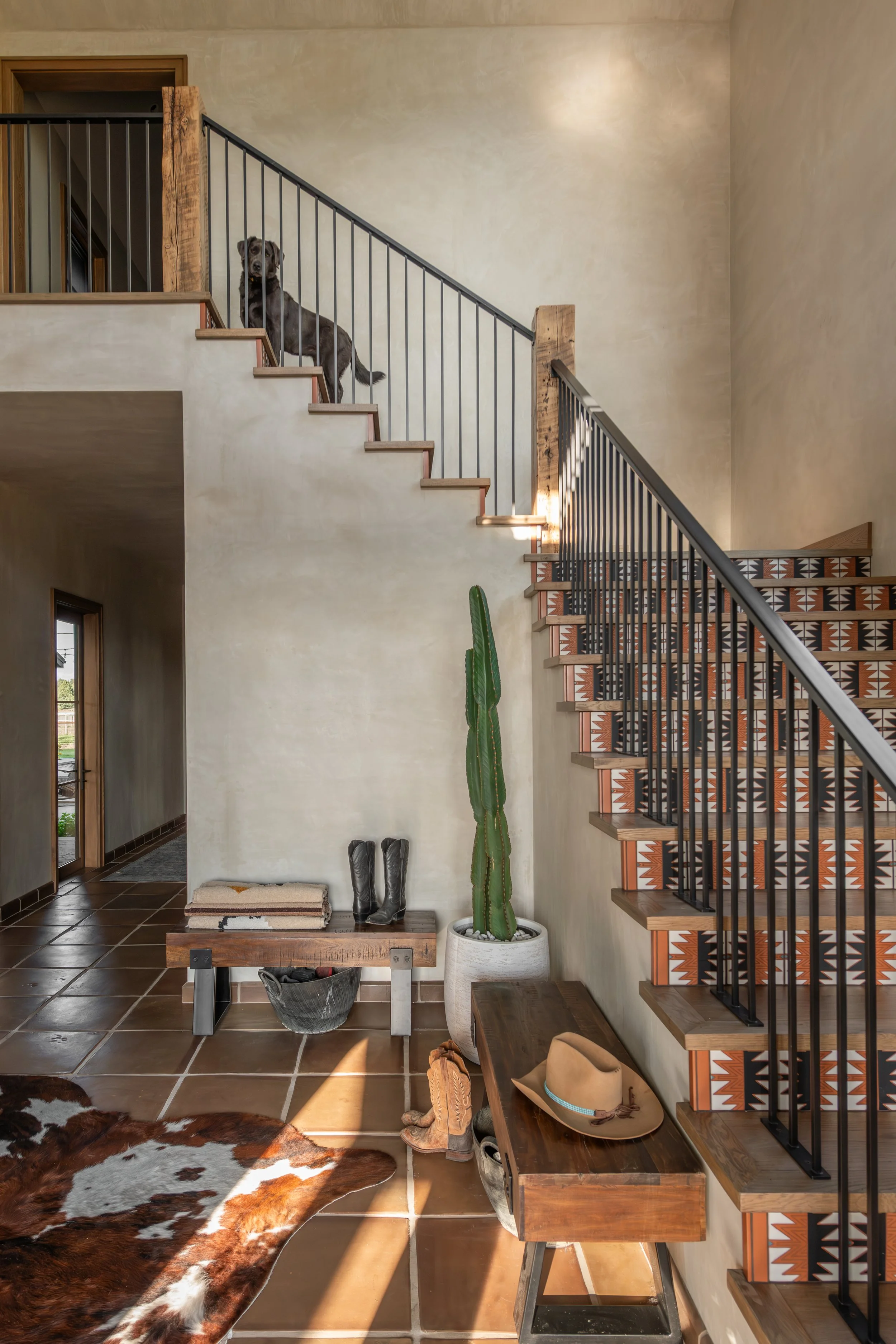 Custom staircase featuring Southwestern patterned tile risers, a black metal railing, and smooth plaster walls in a home built by Copperline Homes in Bend, Oregon. A black dog stands at the top of the stairs.
