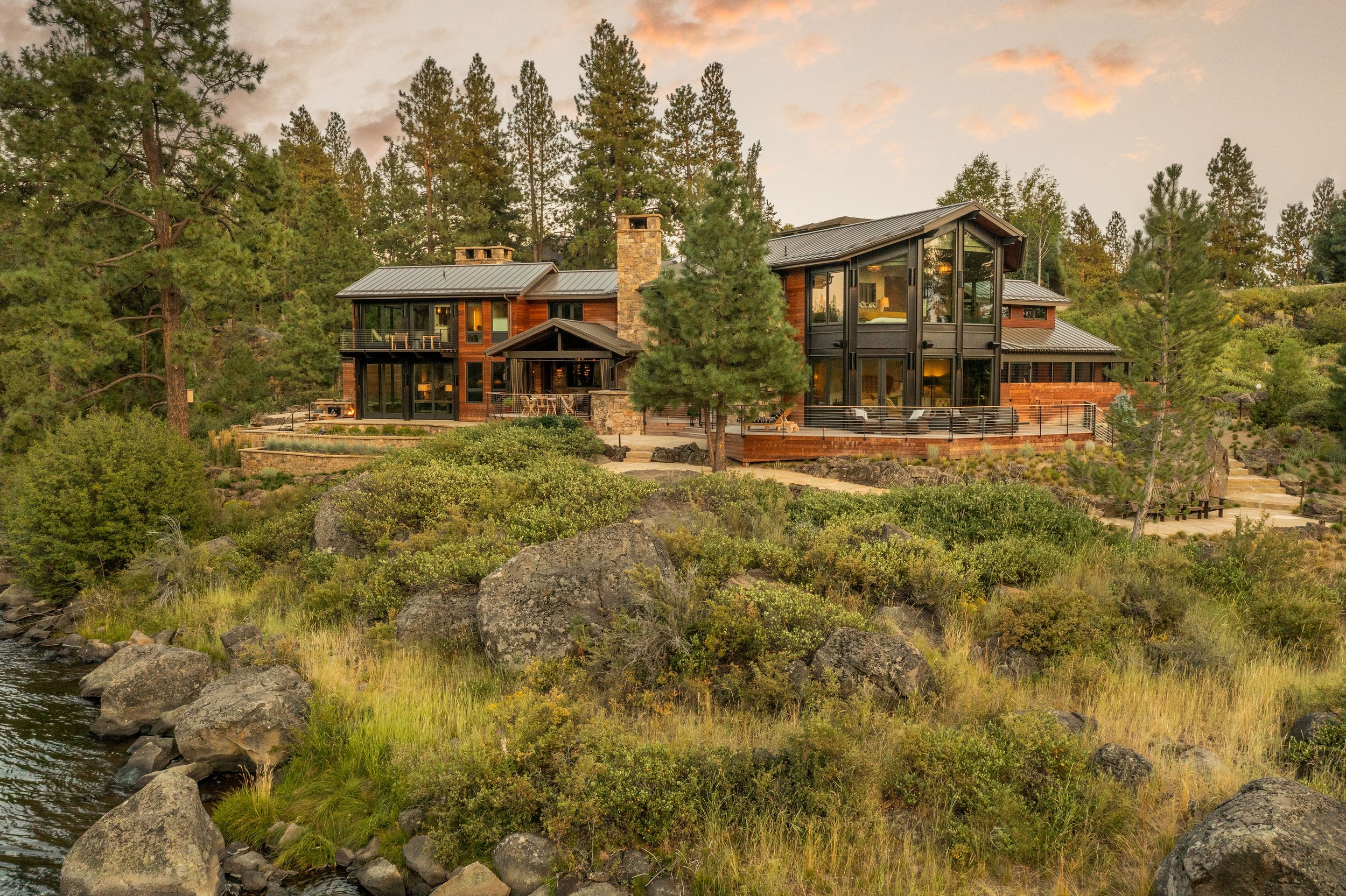 A large modern house with glass windows and a stone chimney, situated on a hillside surrounded by trees and rocks, near a body of water.