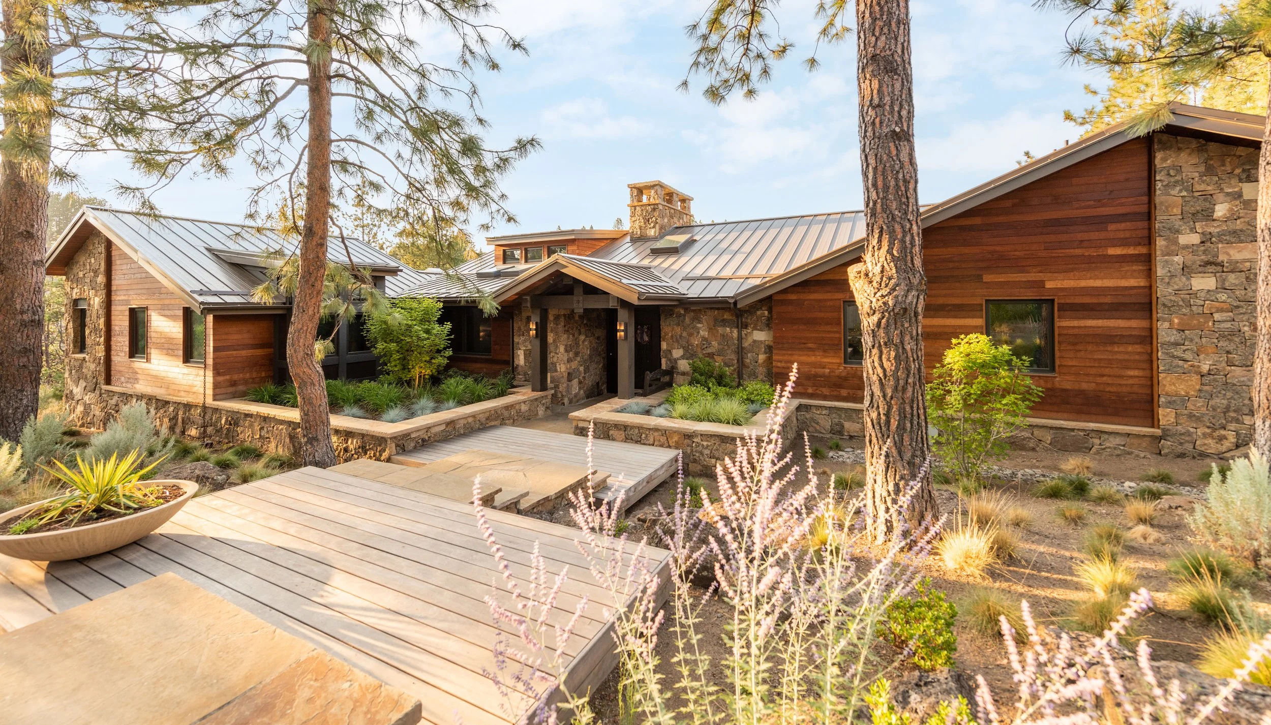 A modern house with a stone and wood exterior, surrounded by tall pine trees and landscaped garden with plants and shrubs, under a blue sky.