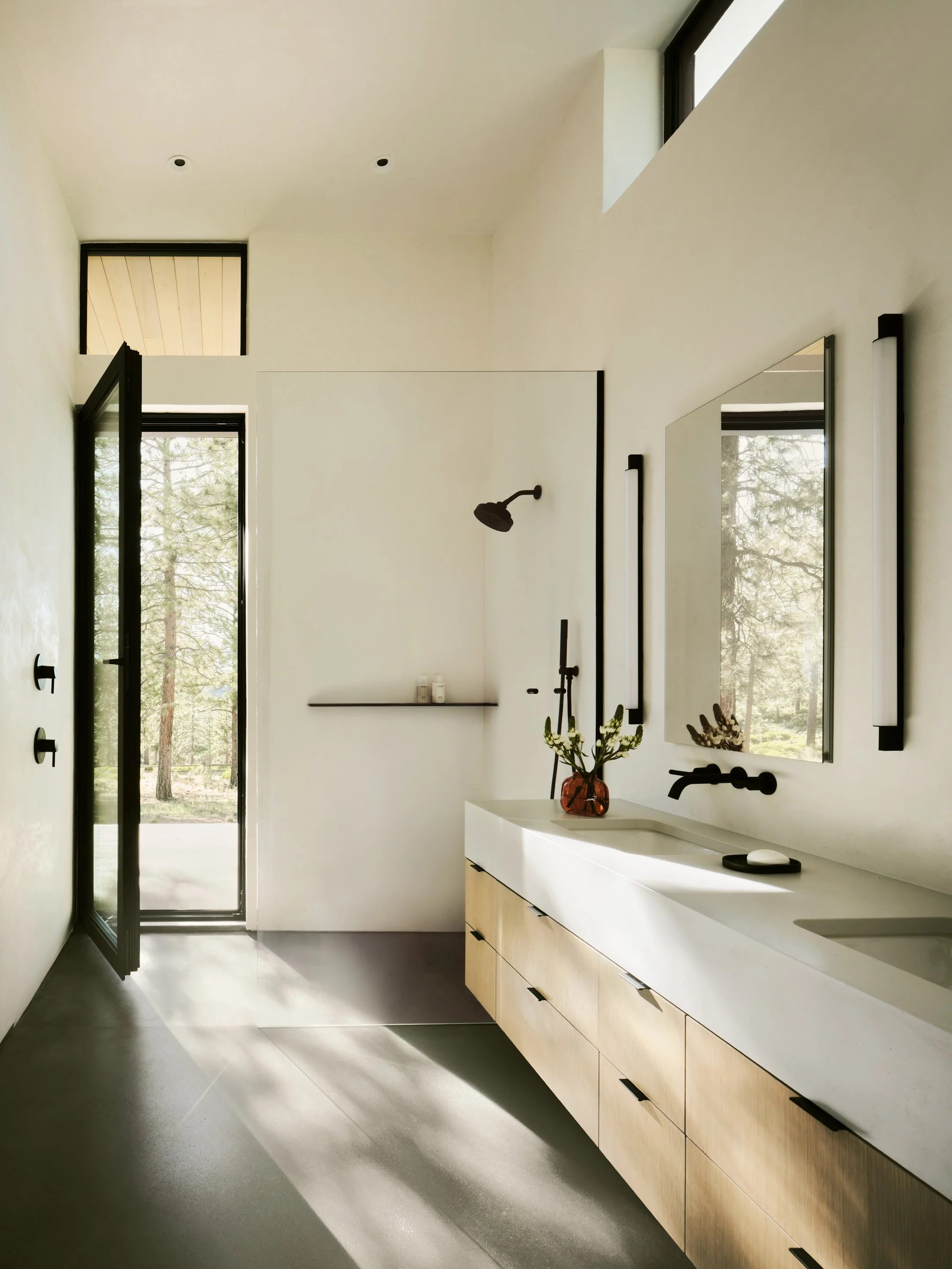 Minimalist bathroom with large mirror, black fixtures, and wooden vanity, featuring a walk-in shower and natural light from windows.