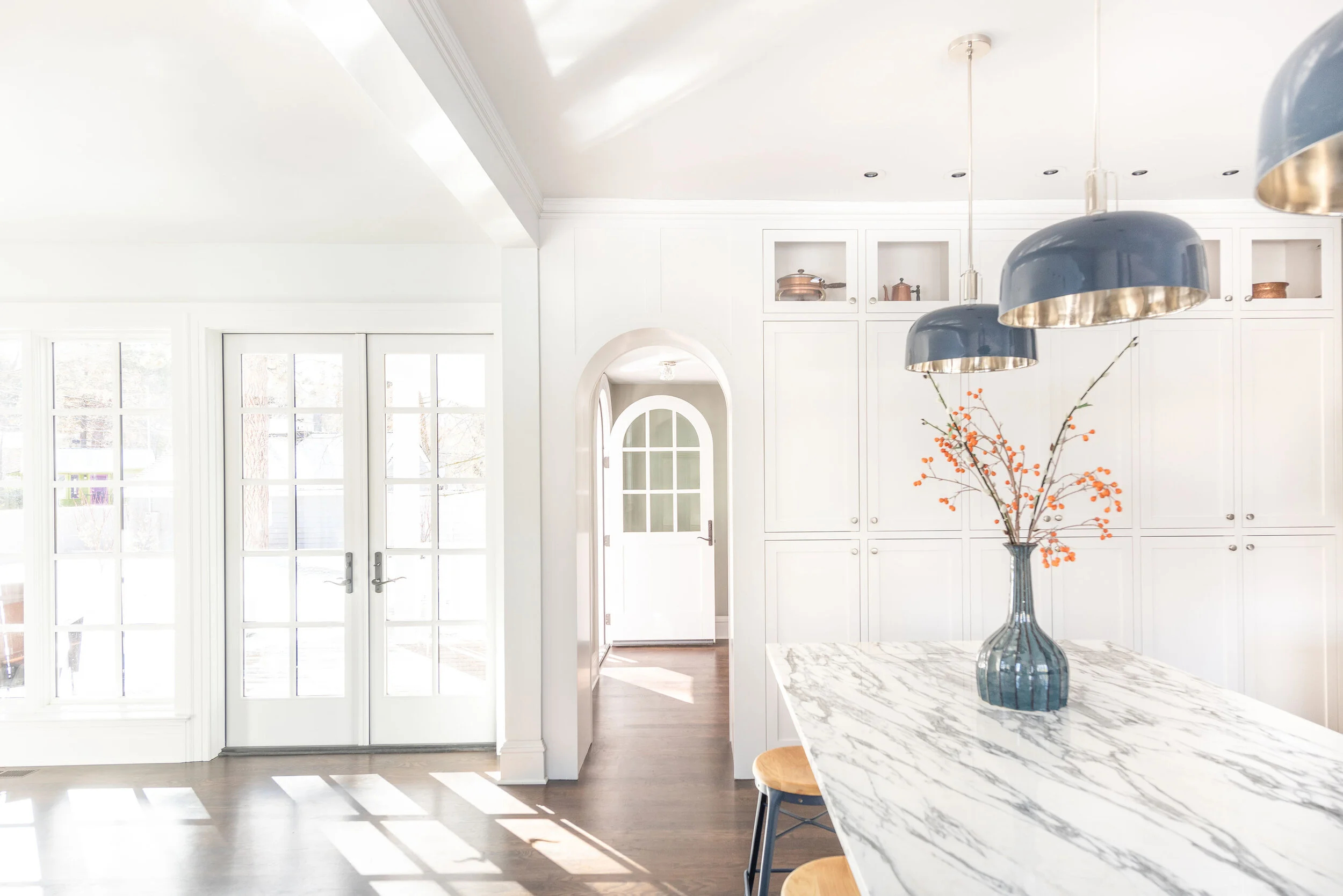 Bright kitchen with white cabinetry and marble countertop island, featuring a vase with orange berries, three large blue pendant lights, and sunlight streaming through glass doors.