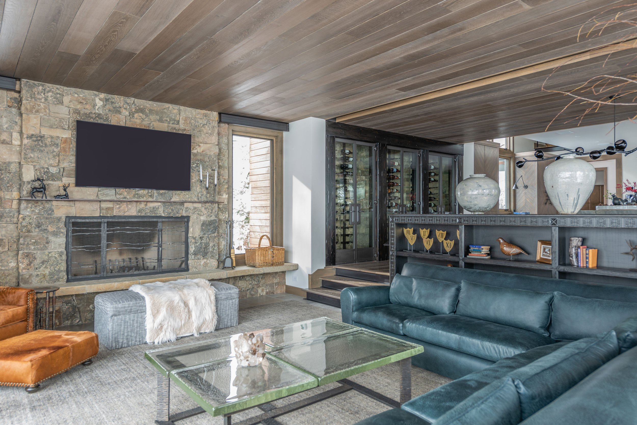 Living room with a stone fireplace, mounted flat-screen TV, leather sofas, glass coffee table, wooden ceiling, large vases, and built-in shelves with decorative items.