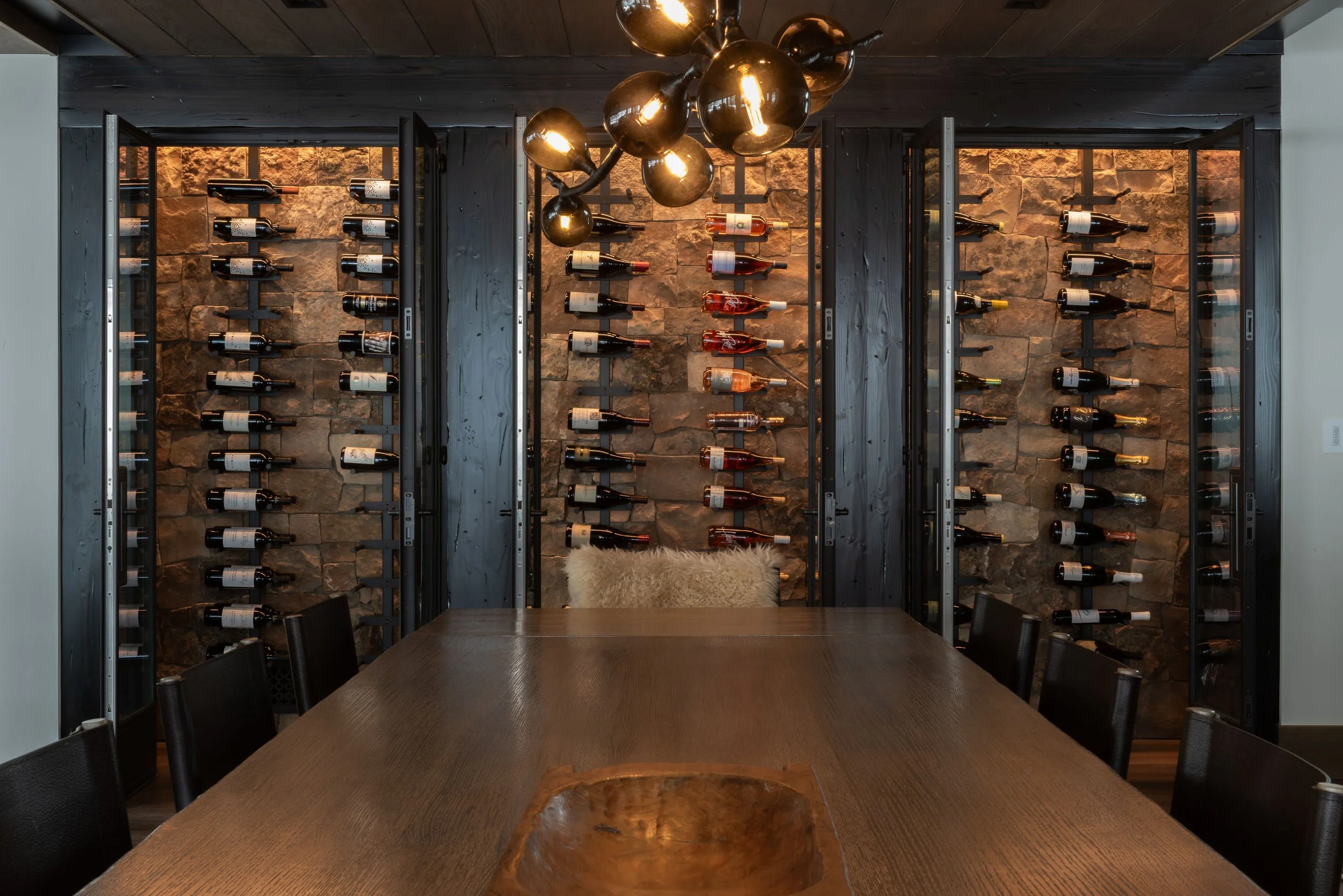 Interior of a dining room with a large wooden table, black chairs, and a wall with a wine rack holding multiple bottles, illuminated by a modern chandelier.