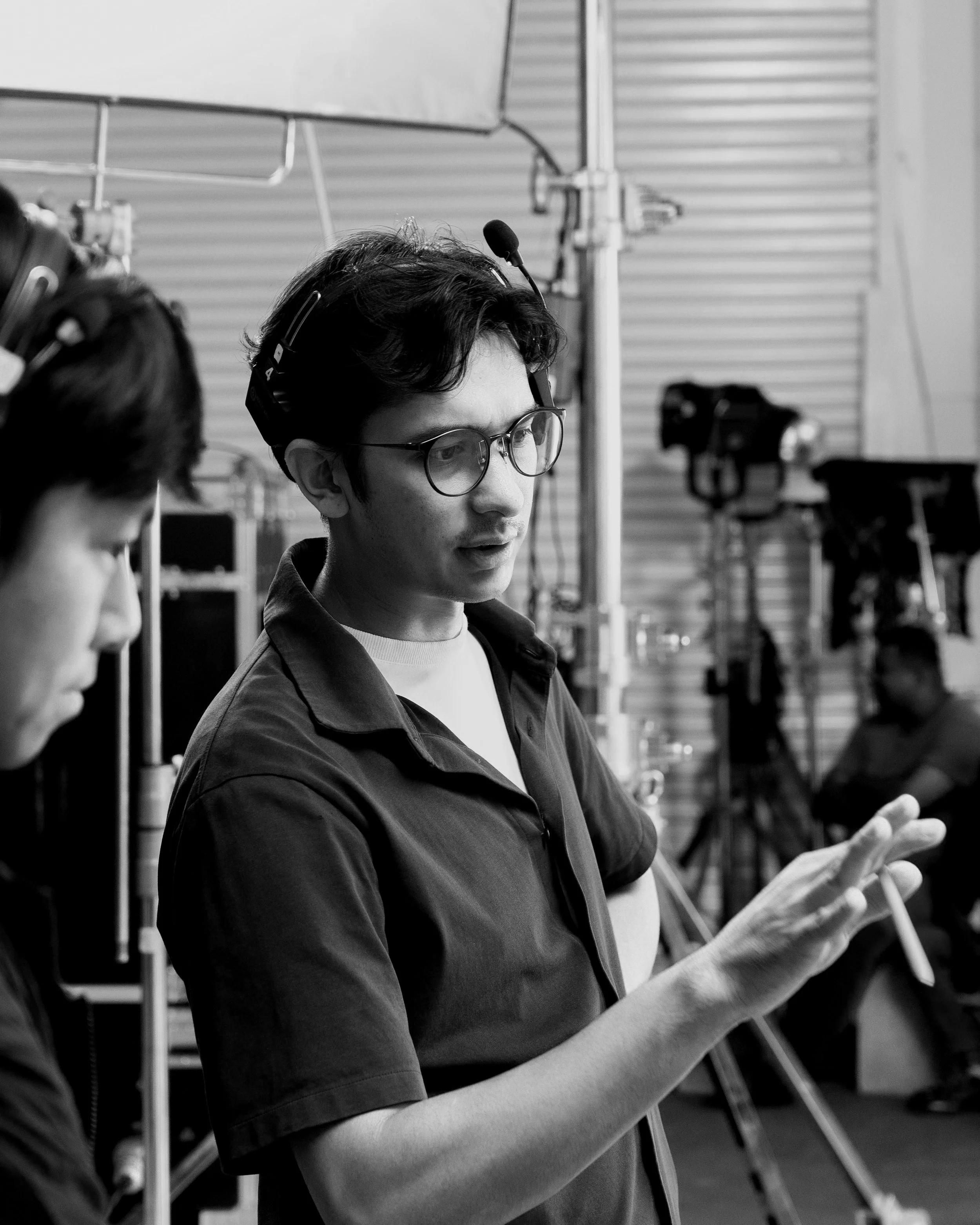 A person wearing glasses and a headset, holding a phone or similar device, seen in a studio or production setting with equipment and crew members in the background.