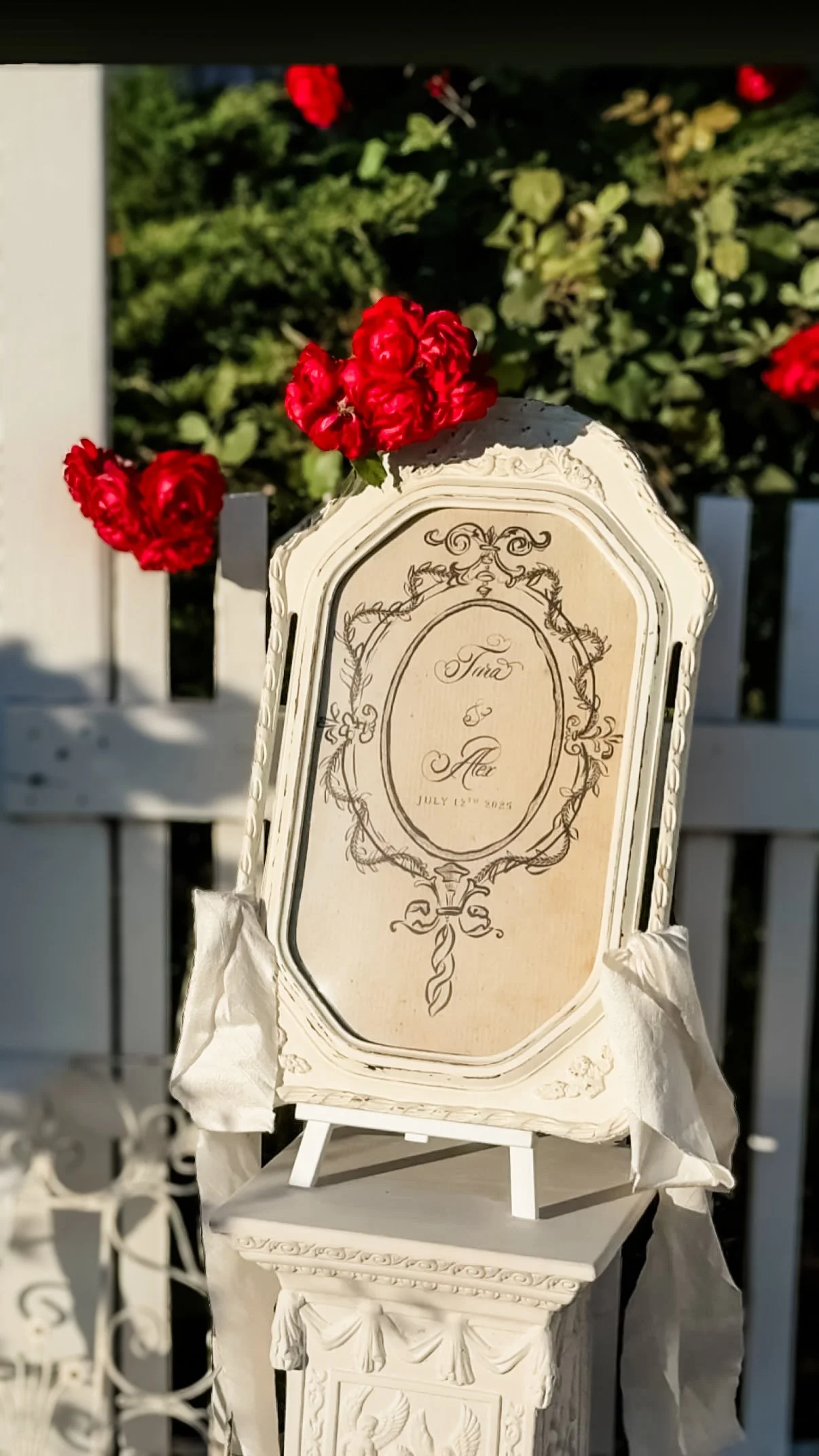 A decorative sign with the names Tara and Alex and the date July 12th, 2023, surrounded by red flowers, displayed on a white pedestal outdoors.