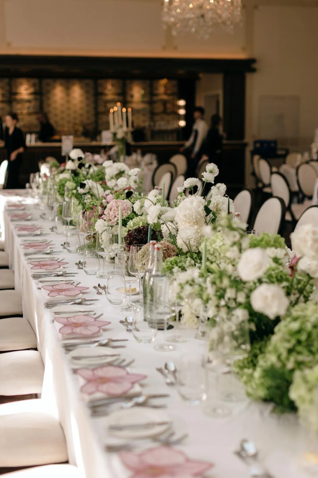 A long banquet table decorated with white and pink flowers, candles, and elegant place settings in a formal event hall.