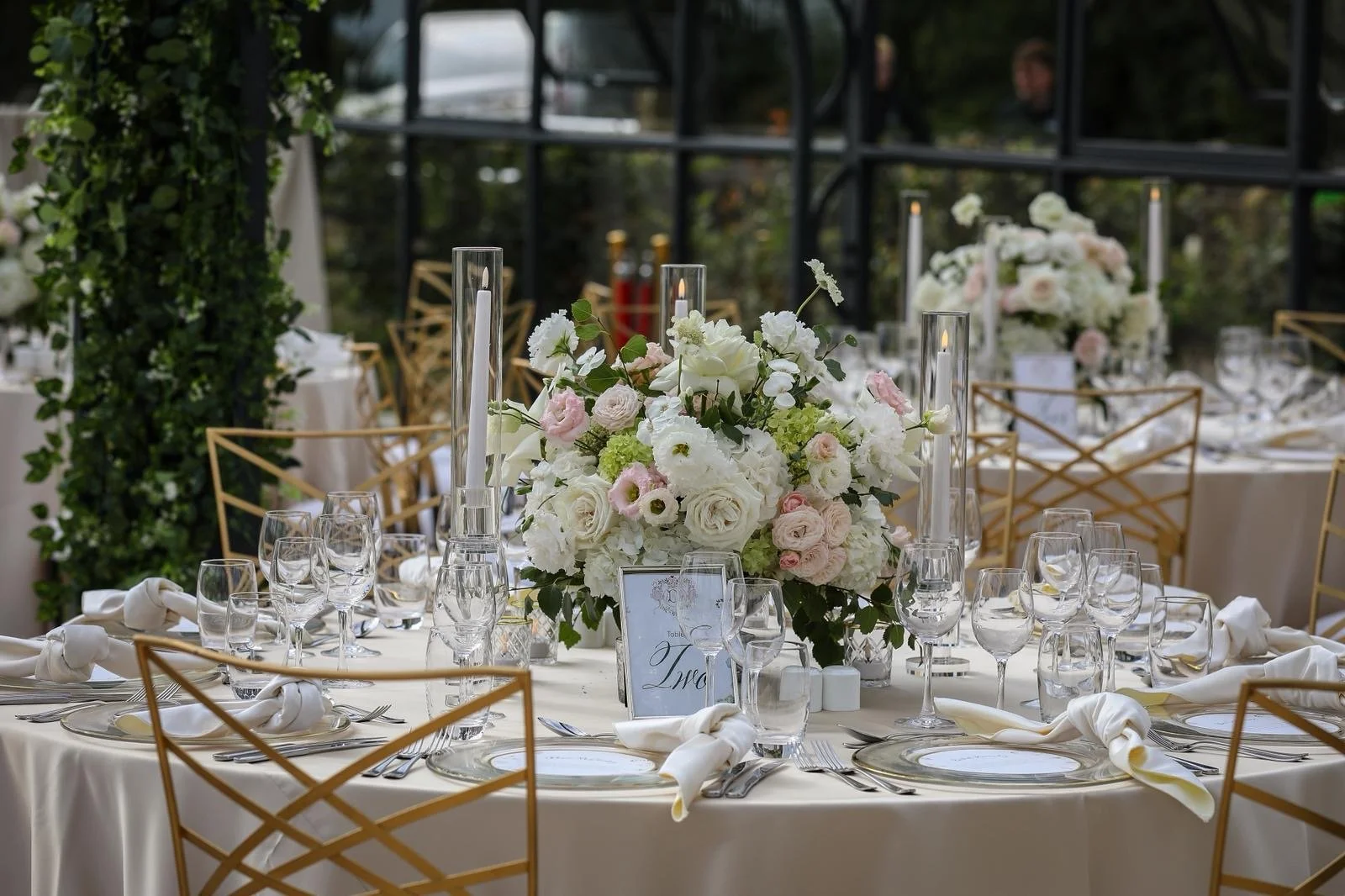 Wedding reception table decorated with a large centerpiece of white and pale pink flowers, tall glass candleholders with lit candles, gold chairs, and elegant table settings including glassware and white napkins, in a bright outdoor venue with greene