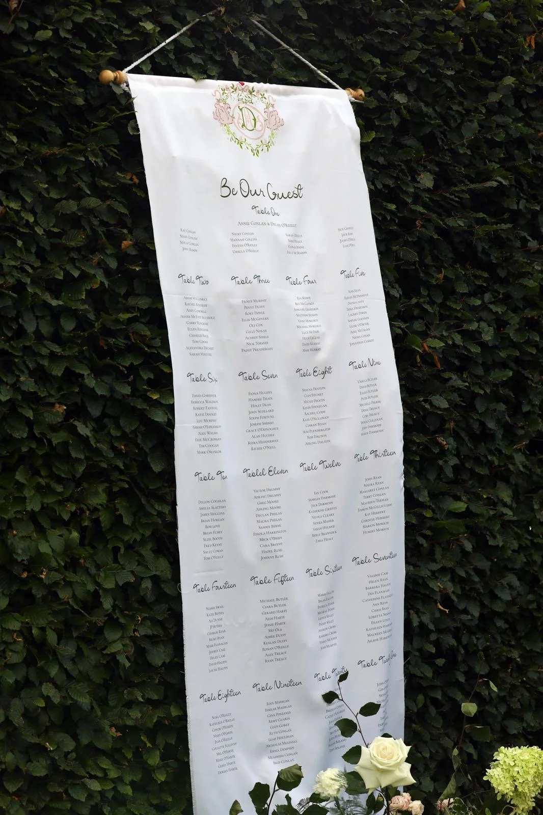 Wedding seating chart displayed on a white fabric banner with calligraphy, hanging against a green leafy background, decorated with white roses and greenery at the bottom.