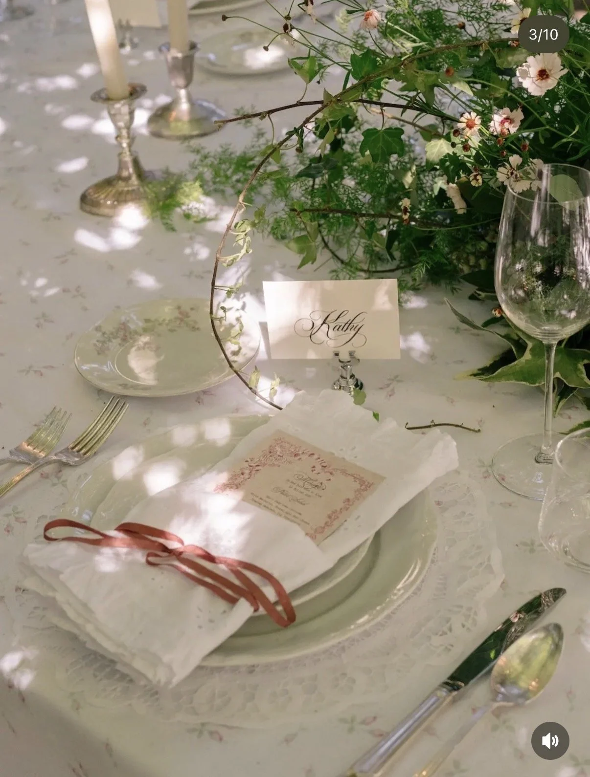 A table setting for a formal event with a white lace tablecloth, a plate stack, a cloth napkin tied with a red ribbon, a printed menu, a small card with the name 'Kaitty', greenery and flowers as a centerpiece, and candles in the background.