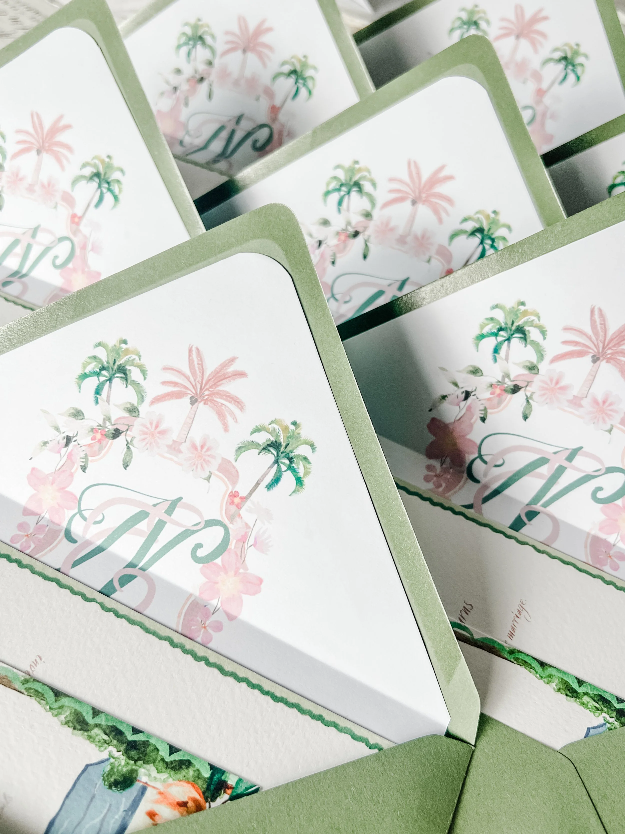 Multiple green and white cards with tropical designs and initials, decorated with pink and green palm trees and flowers.