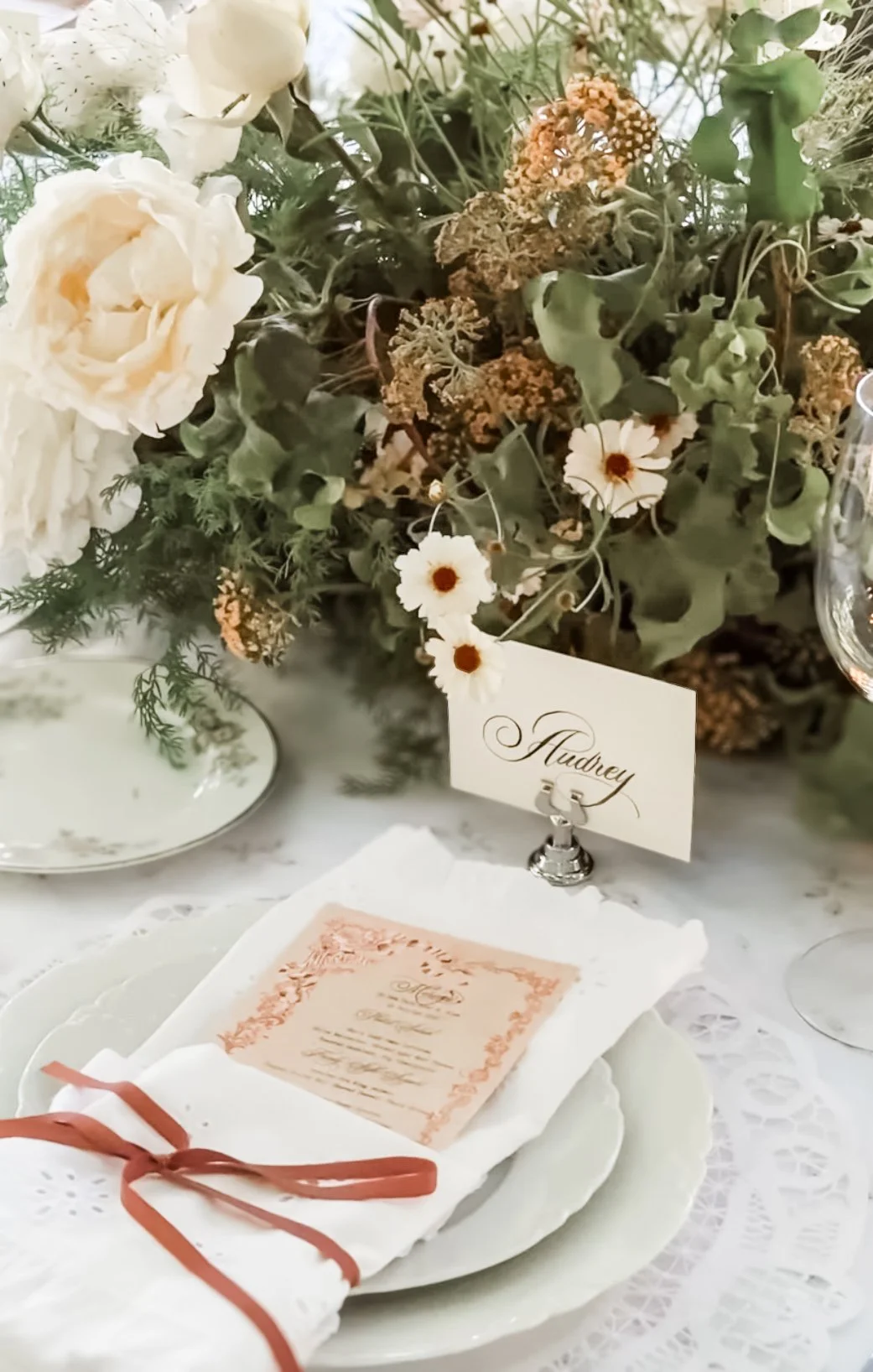 Table setting with a large floral centerpiece, place card labeled 'Audrey,' and a menu wrapped in a napkin with a brown ribbon, on a lace tablecloth.