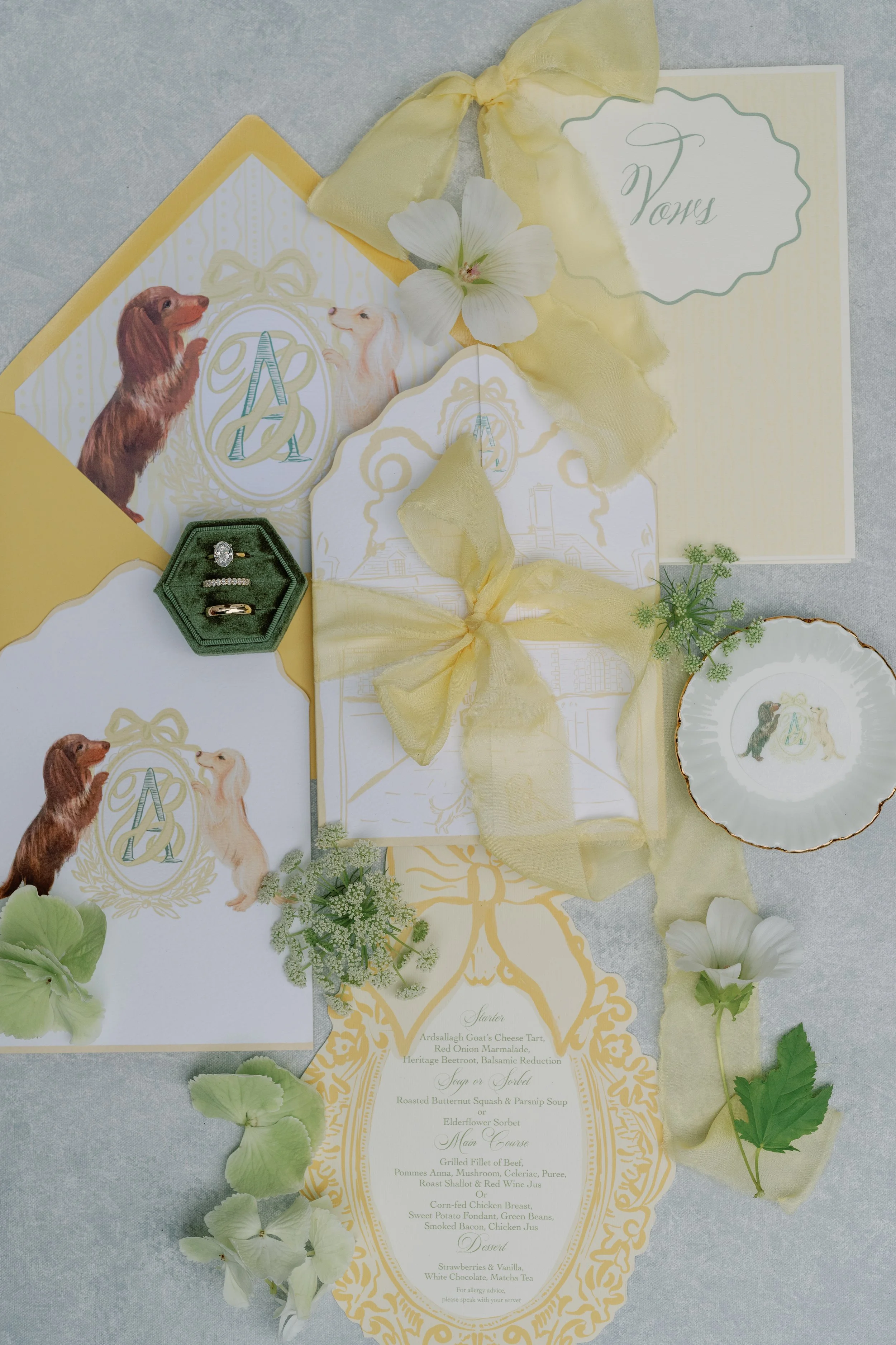 Elegant wedding stationery display with illustrated dog and cat animals, yellow ribbons, green foliage, a jewelry ring, and a menu card with gold ornate design.