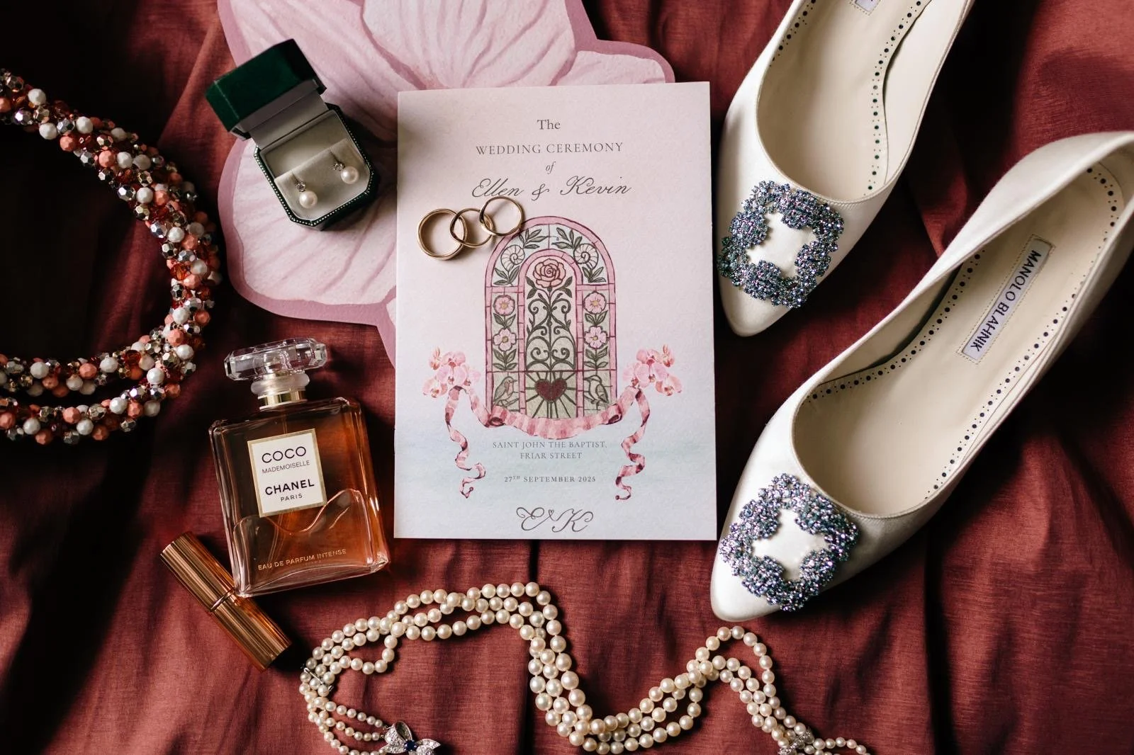 Wedding invitation surrounded by jewelry, shoes, perfume, and accessories on a burgundy fabric background.