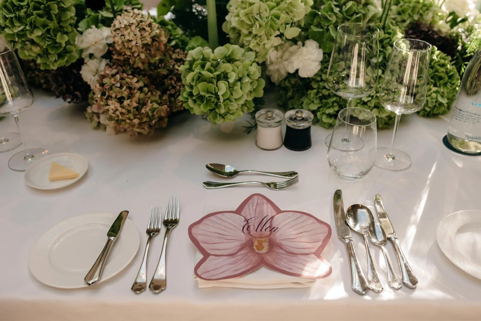 Elegant table setting with floral centerpiece, wine glasses, water glasses, silverware, a plate with a small piece of cheese, and a napkin with the name 'Ellen' on a flower-shaped paper. Salt and pepper shakers are also on the table.