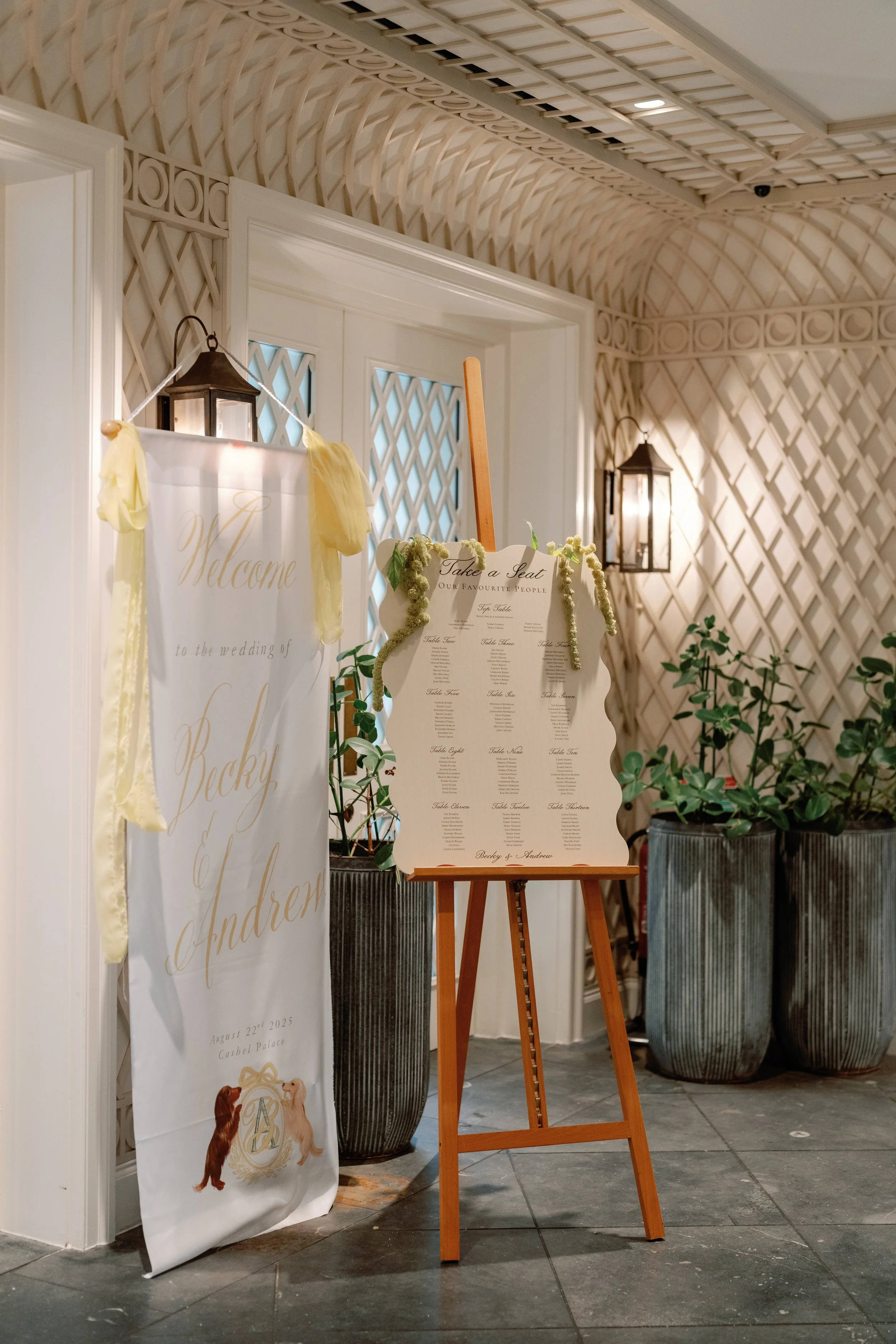 Wedding reception area with a seating chart on an easel and a welcome sign with floral decorations, set in an elegant indoor venue with potted plants and decorative wall panels.