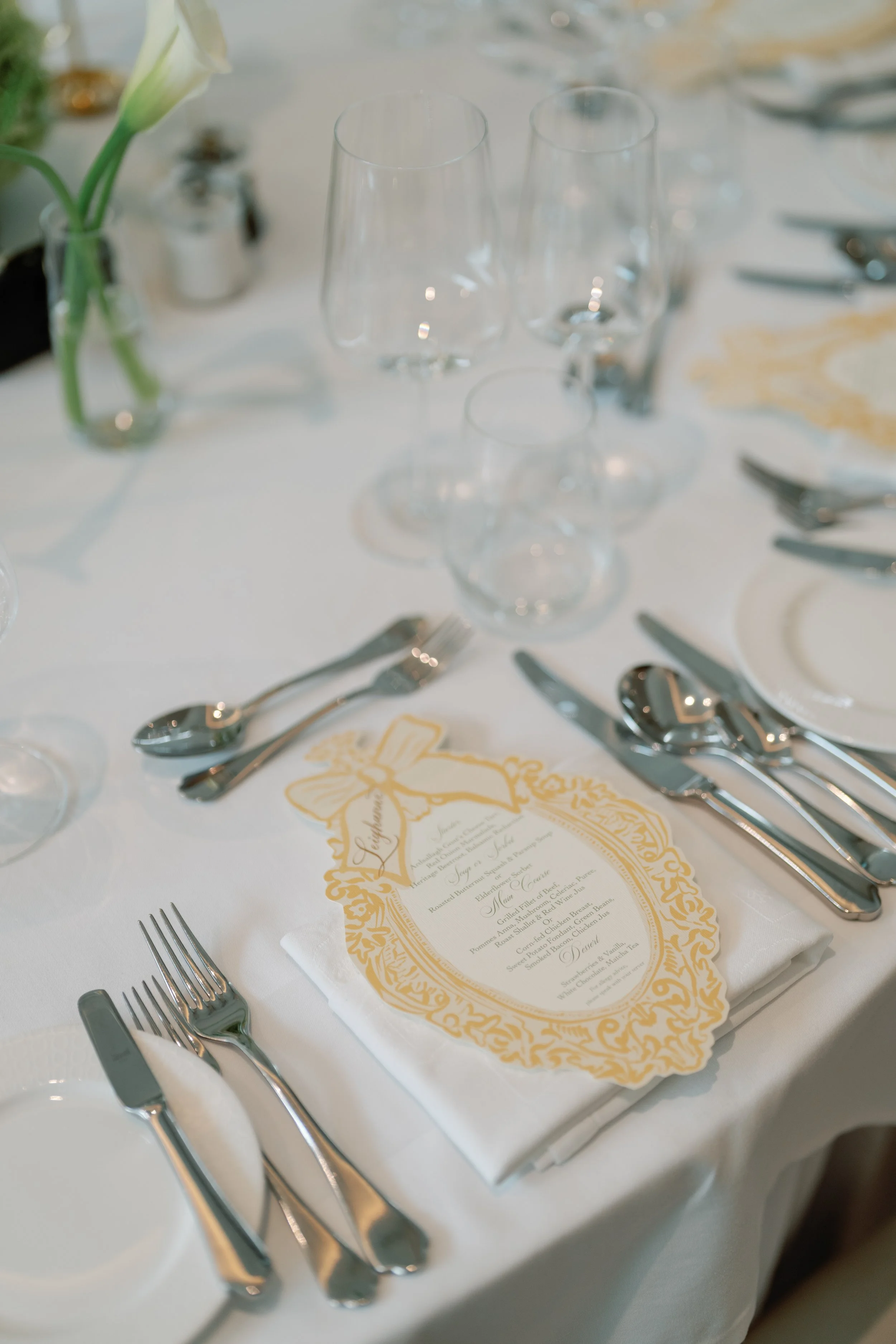 Elegant event table setting with multiple wine glasses, a floral centerpiece, and a printed menu on a white napkin.