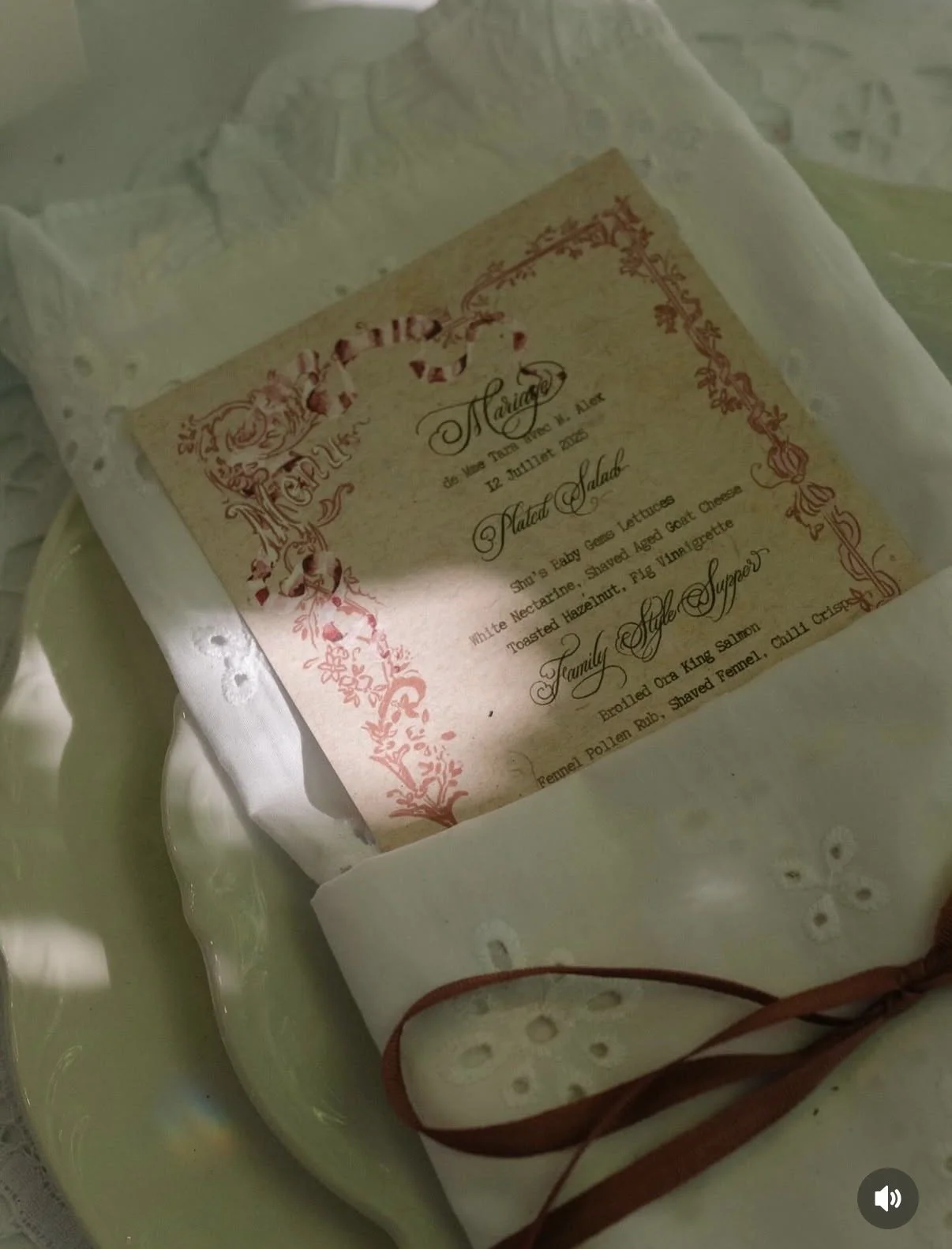 A decorative menu or invitation with ornate pink border and floral design, wrapped in white embroidered fabric with a brown ribbon.