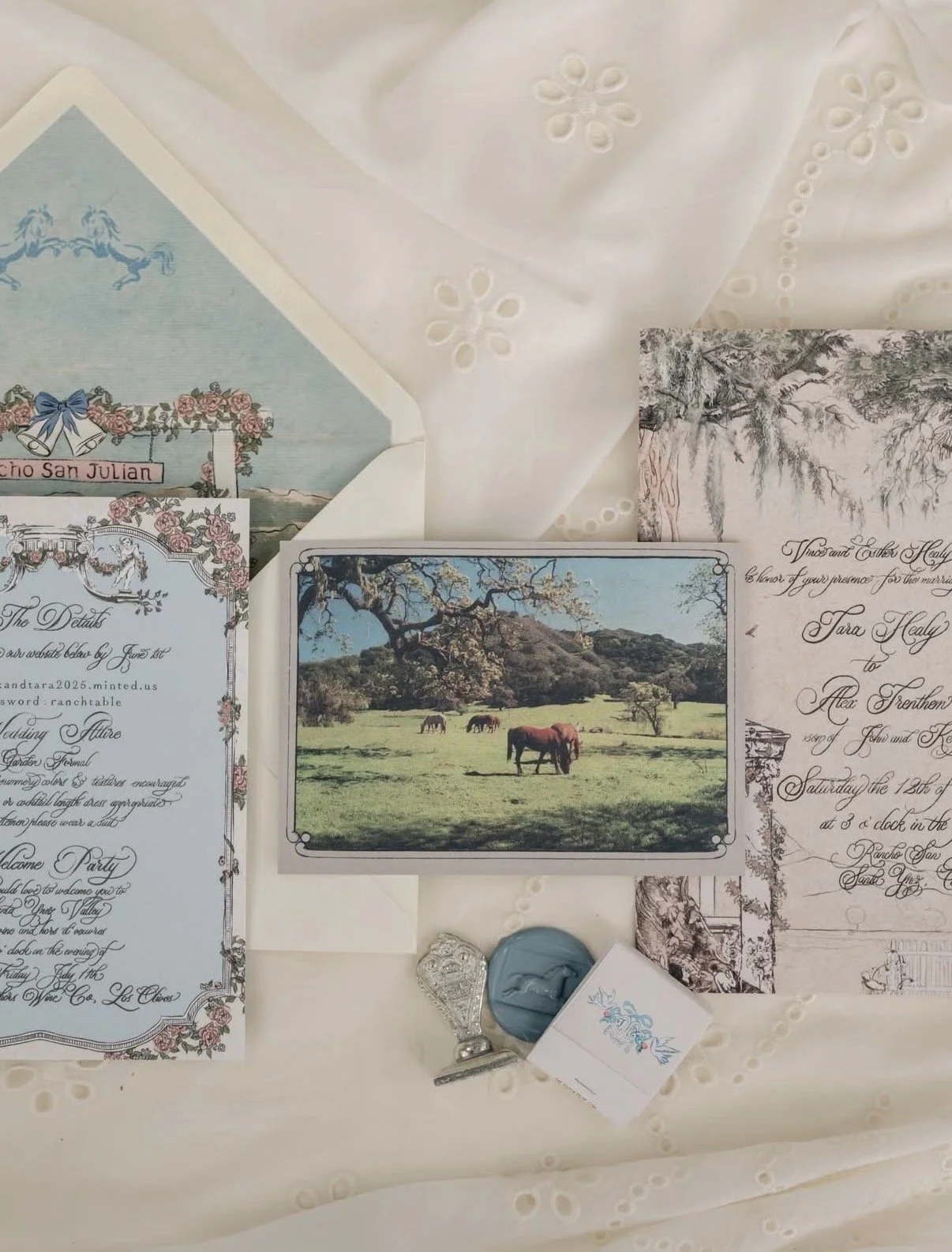 Wedding invitations, a photo of a farm scene with horses, and decorative stamps on a white embroidered fabric.