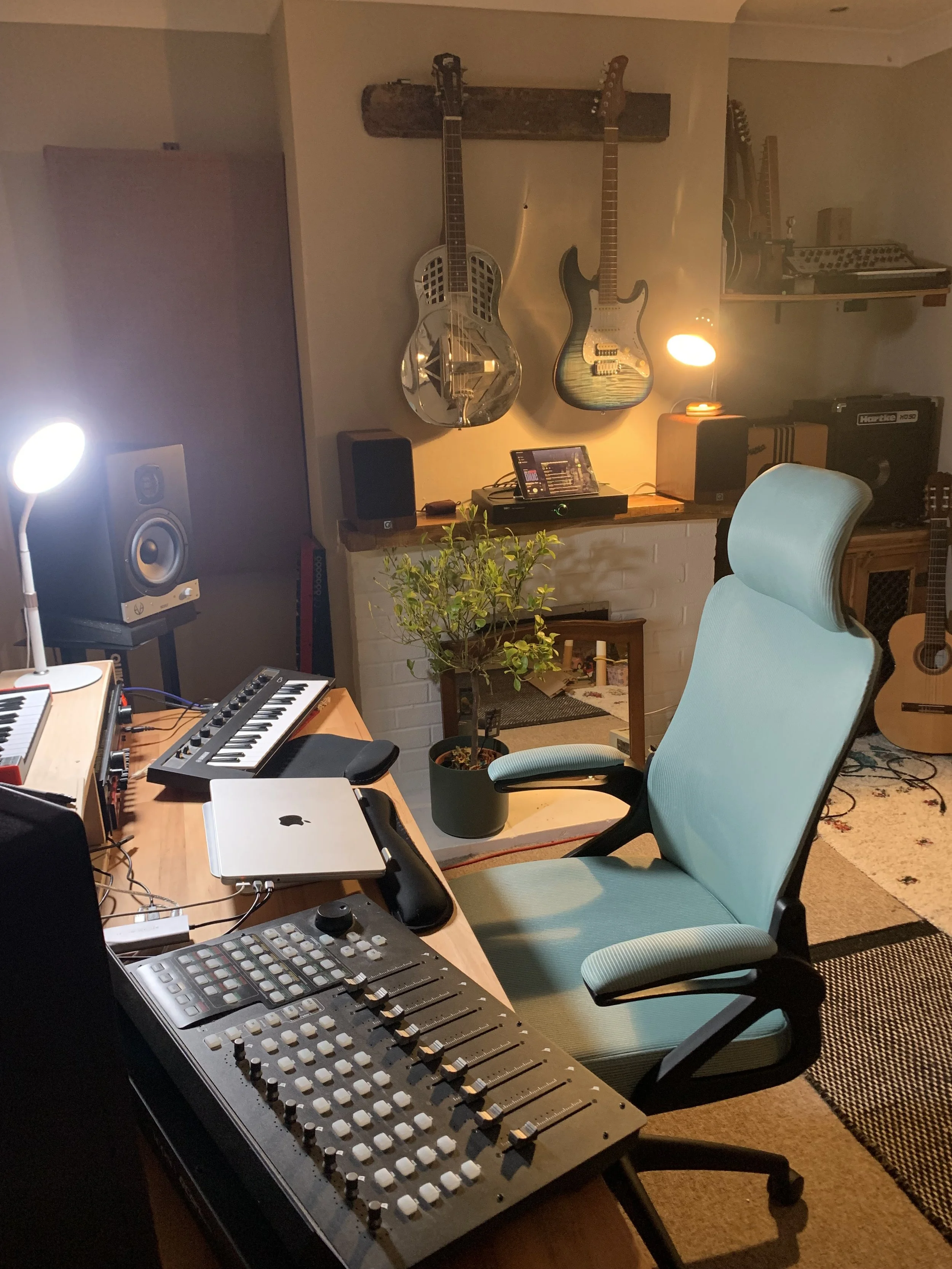 A music studio with a desk containing a mixer, laptop, and keyboard, a light blue ergonomic chair, a small potted plant, and guitars hanging on the wall.
