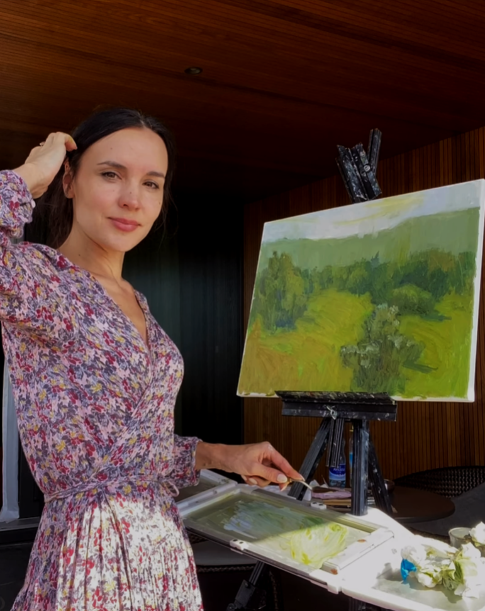 Eine Frau in einem bunten Kleid steht vor einer Staffelei und malt eine Landschaft auf Leinwand. Sie berührt ihr Haar mit einer Hand.