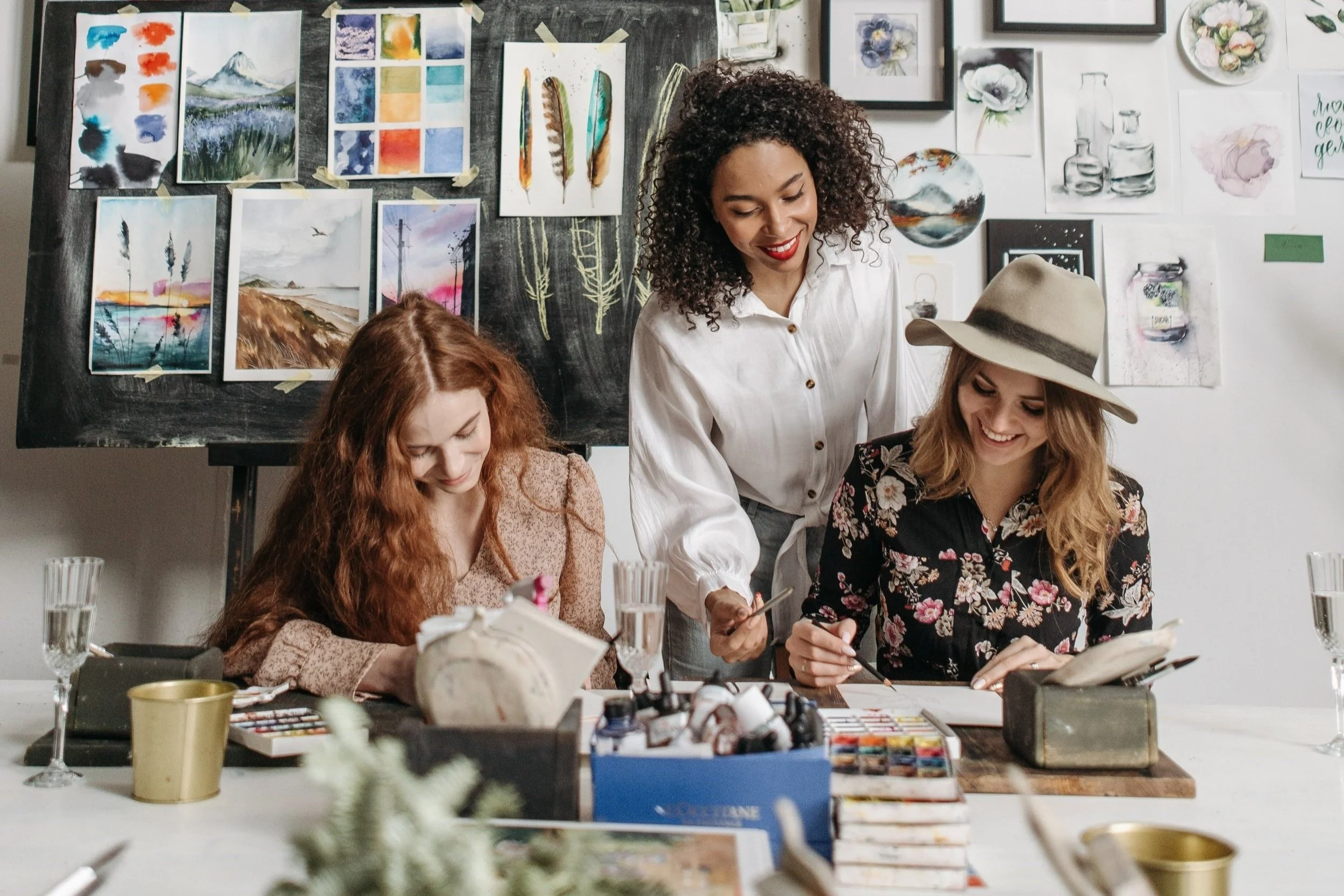 Drei Frauen beim Malen und Skizzieren in einem Kunststudio. Im Hintergrund sind Wasserfarben, Aquarellbilder und Skizzen an der Wand. Es gibt Pinsel, Farben, Skizzenbücher und Wasser gefüllte Gläser auf dem Tisch.