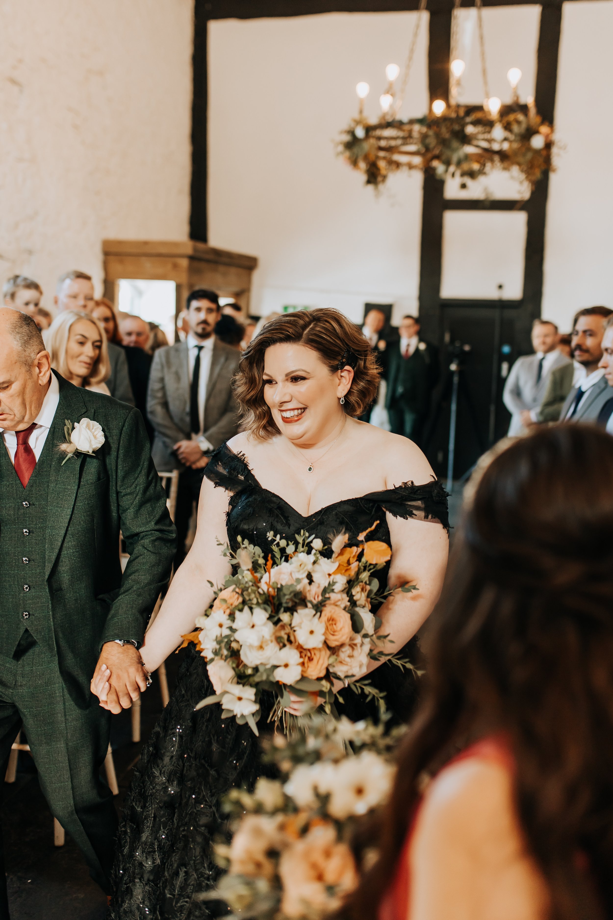 A bride in a black dress holding a bouquet, walking down the aisle, accompanied by a man in a suit. Guests are smiling and watching the ceremony inside a decorated venue with a chandelier overhead.