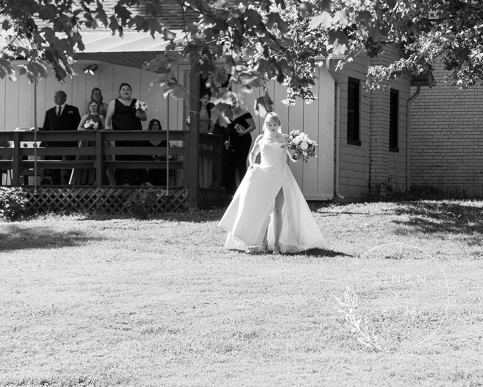 A bride holding a bouquet walks on a grassy yard, with bridesmaids and a man standing behind on a porch in the background. The image is in black and white.