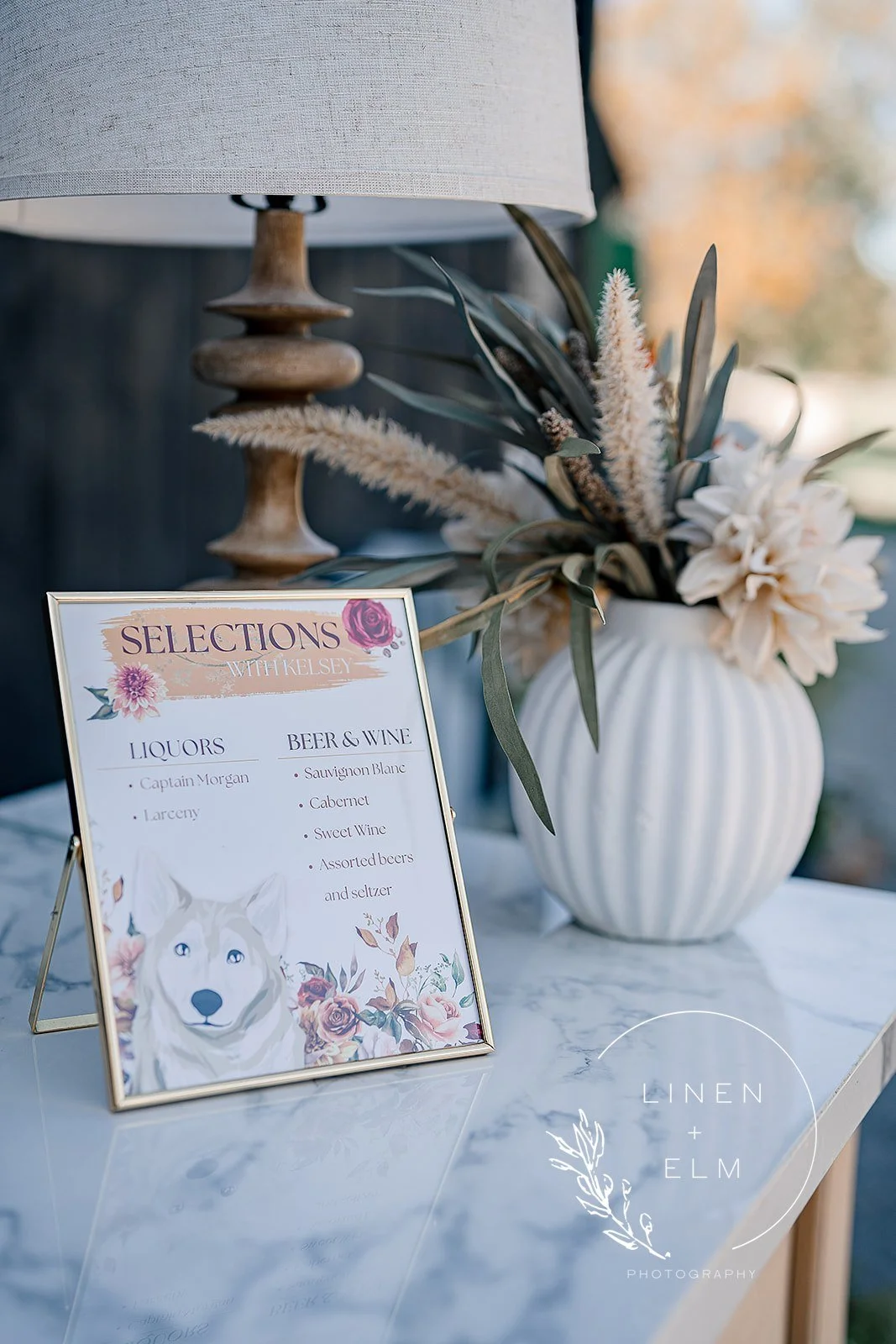 A table with a framed sign listing drink options, a white vase with dried flowers, and a wooden lamp, with an outdoor background.