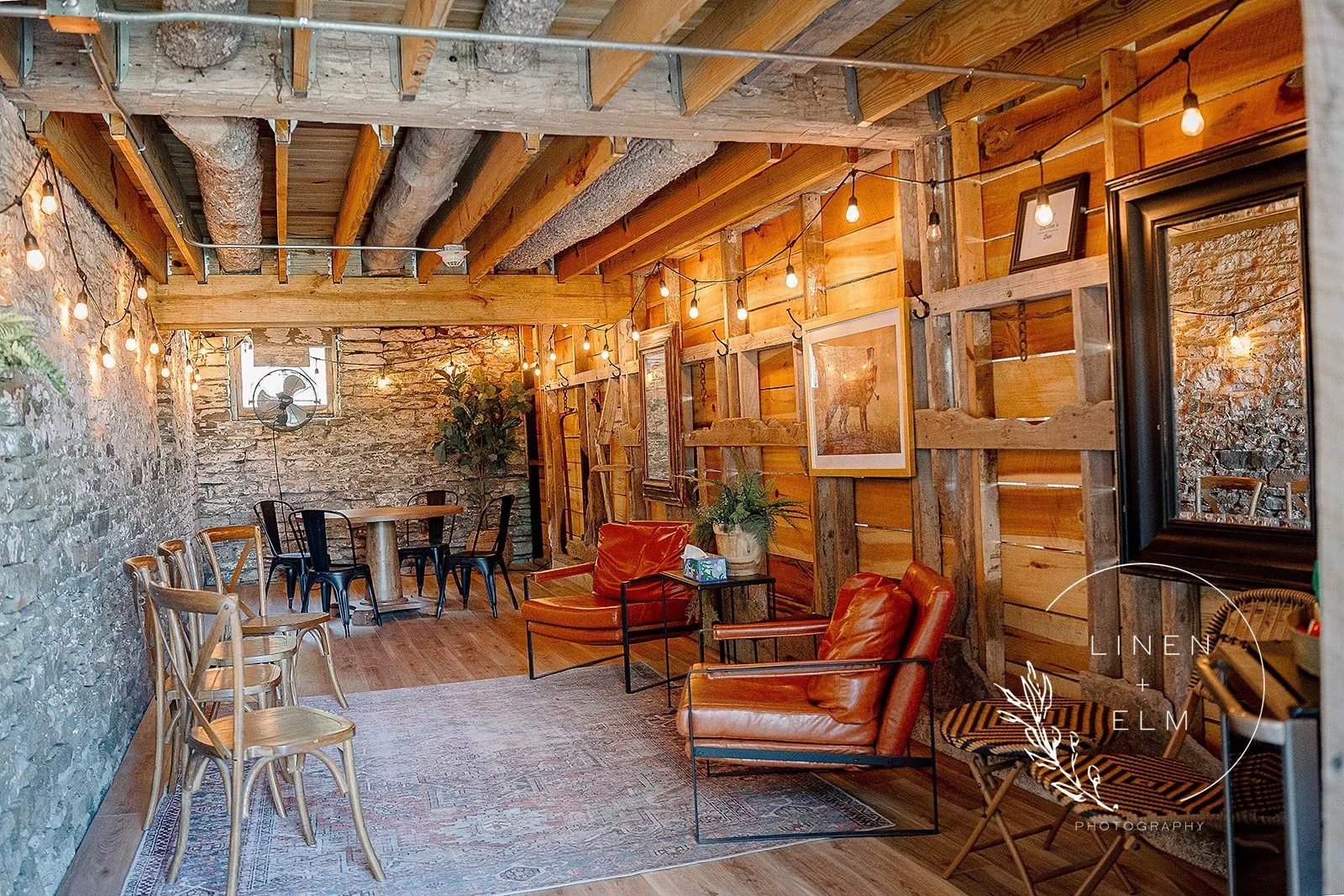 Cozy interior of a rustic-style room with exposed stone and wood walls, string lights hanging from the ceiling, leather armchairs, framed artwork, potted plants, and a small round dining table with chairs.