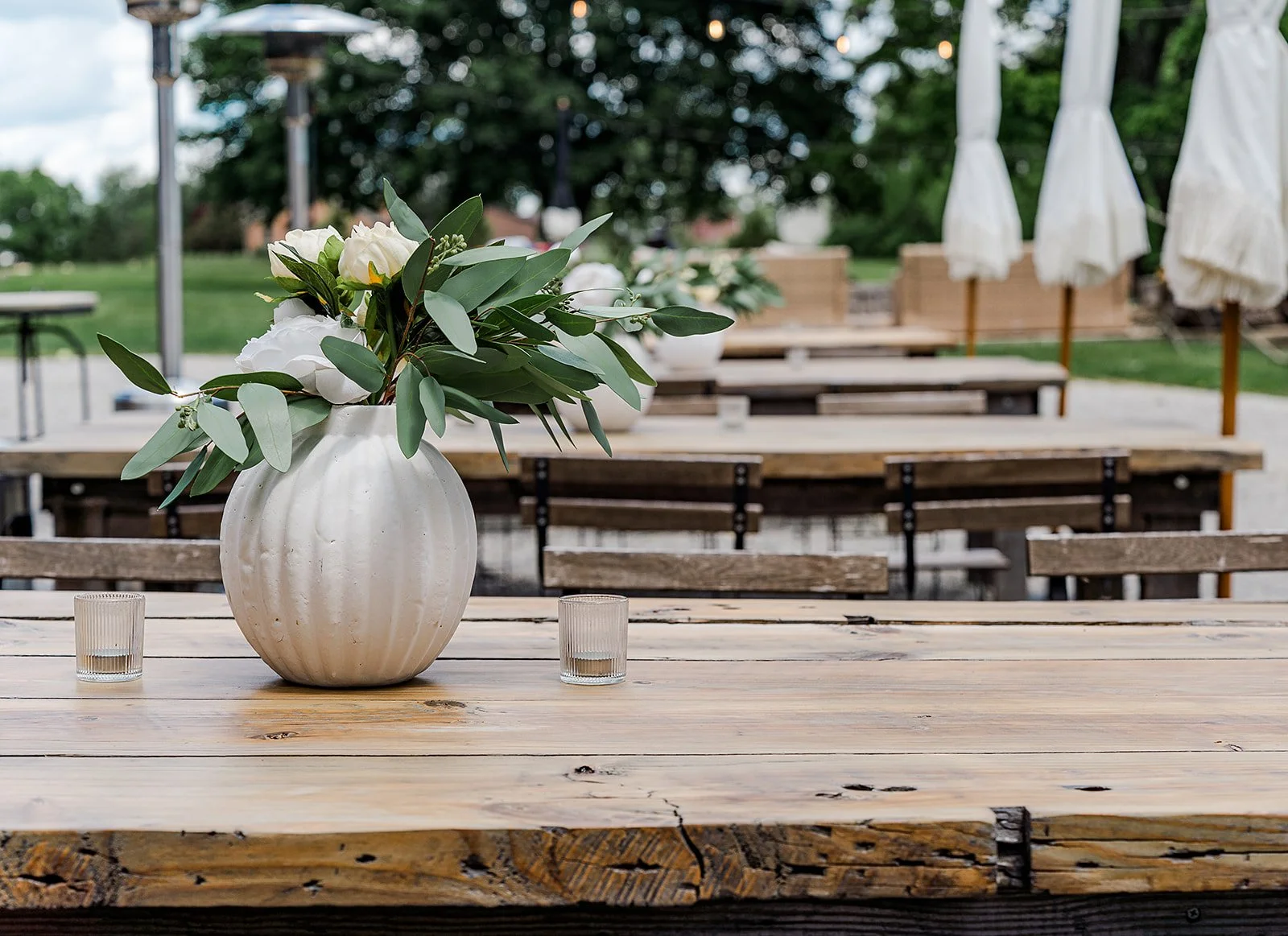 A white ceramic vase with green leaves and white flowers on a wooden table, two small glass candles nearby, outdoor patio setting with wooden benches and white umbrellas in the background at a 19th century estate