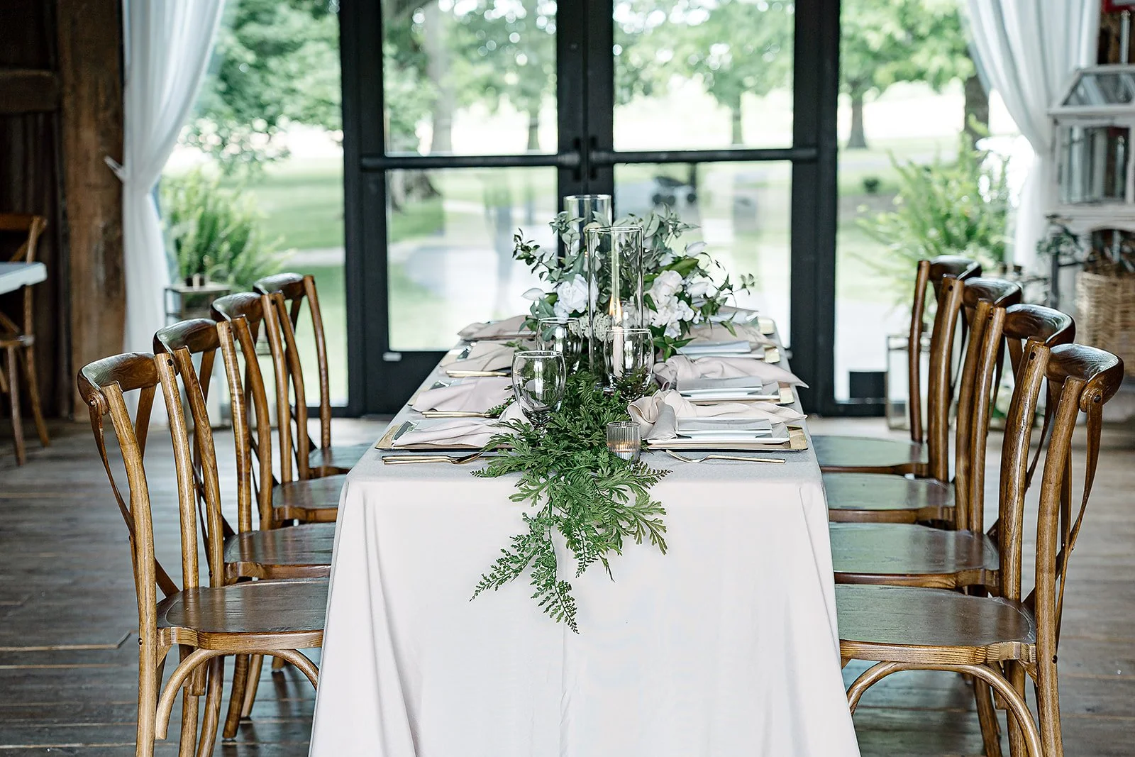 Elegant wedding or event table setup with white tablecloth, floral centerpiece, greenery, candle, glassware, and silverware surrounded by wooden chairs inside a rustic venue with large glass doors and view of greenery outside.