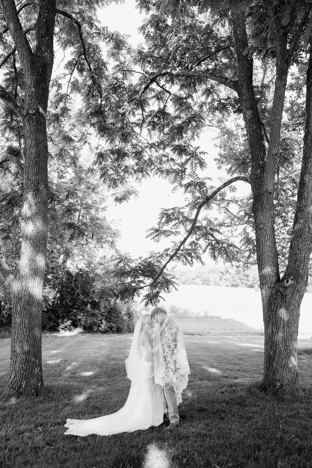 A black and white photo of a couple in wedding attire standing under tall trees in a grassy field, sharing a moment of intimacy at a 19th century estate