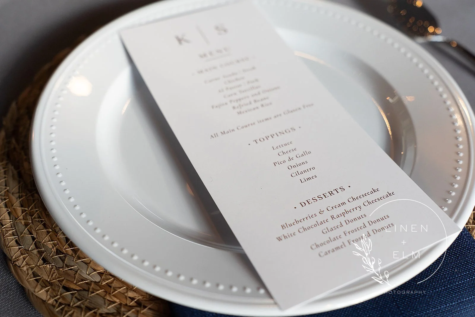 Close-up of a wedding menu on white paper, placed on a white ceramic plate with a decorative dotted border, set on a woven placemat. The menu lists food and dessert options.