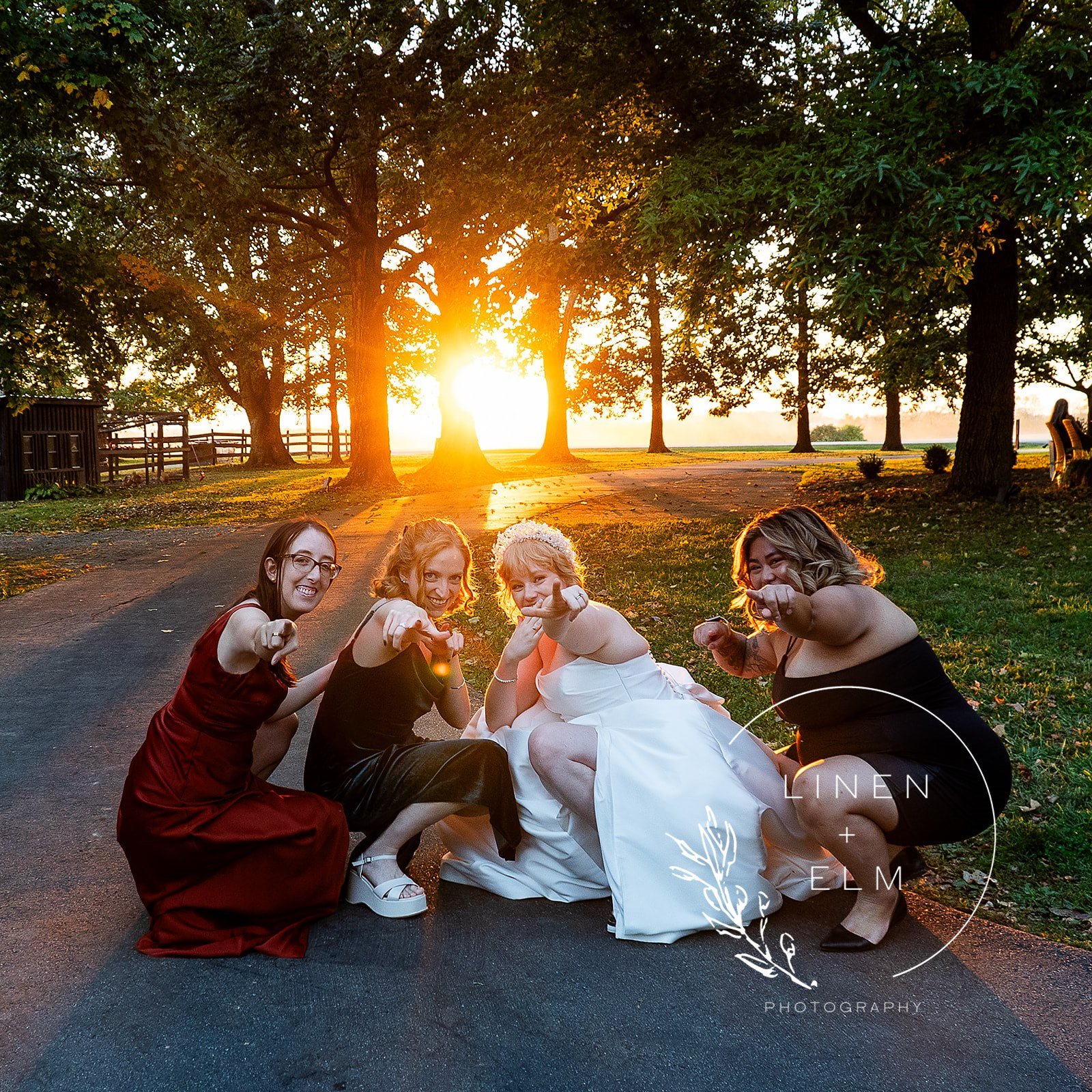 Group of four women in formal dresses posing together outdoors at sunset, with trees and a pathway in the background.