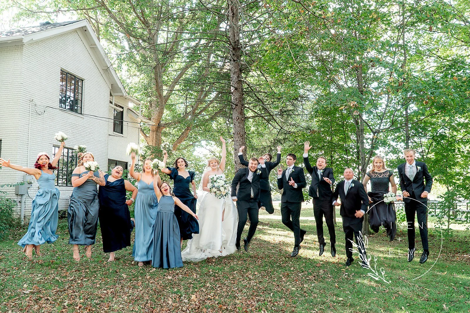 A group of wedding party members, including bridesmaids and groomsmen, celebrating and jumping in the air outdoors on a grassy area with trees and a white brick building in the background.