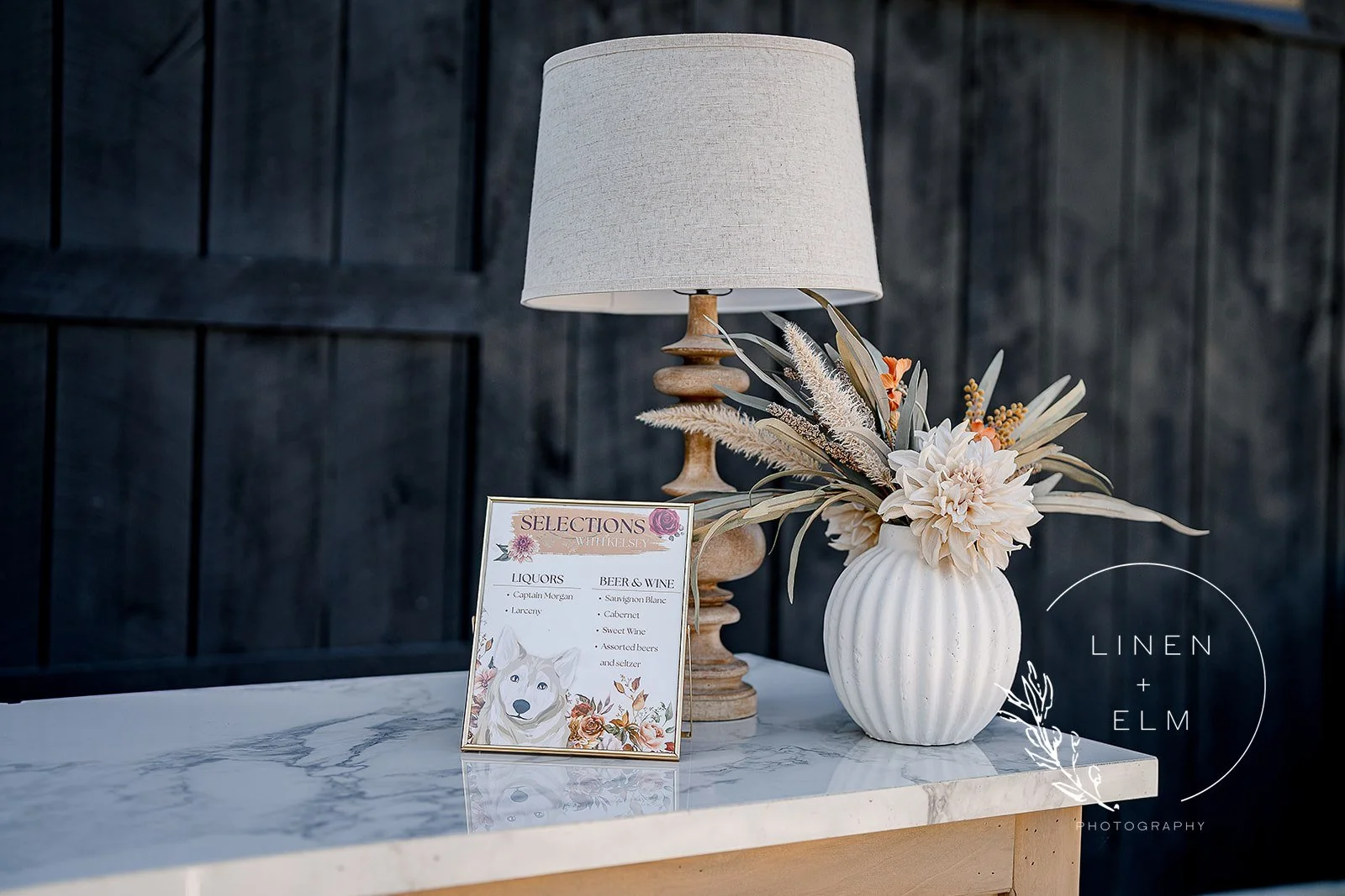 A table with a white marble top, a beige table lamp, a white vase with dried flowers, and a menu card. The wall behind is dark wood paneling, and there is a circular logo with the words "Linen + Elm Photography" in the bottom right corner.