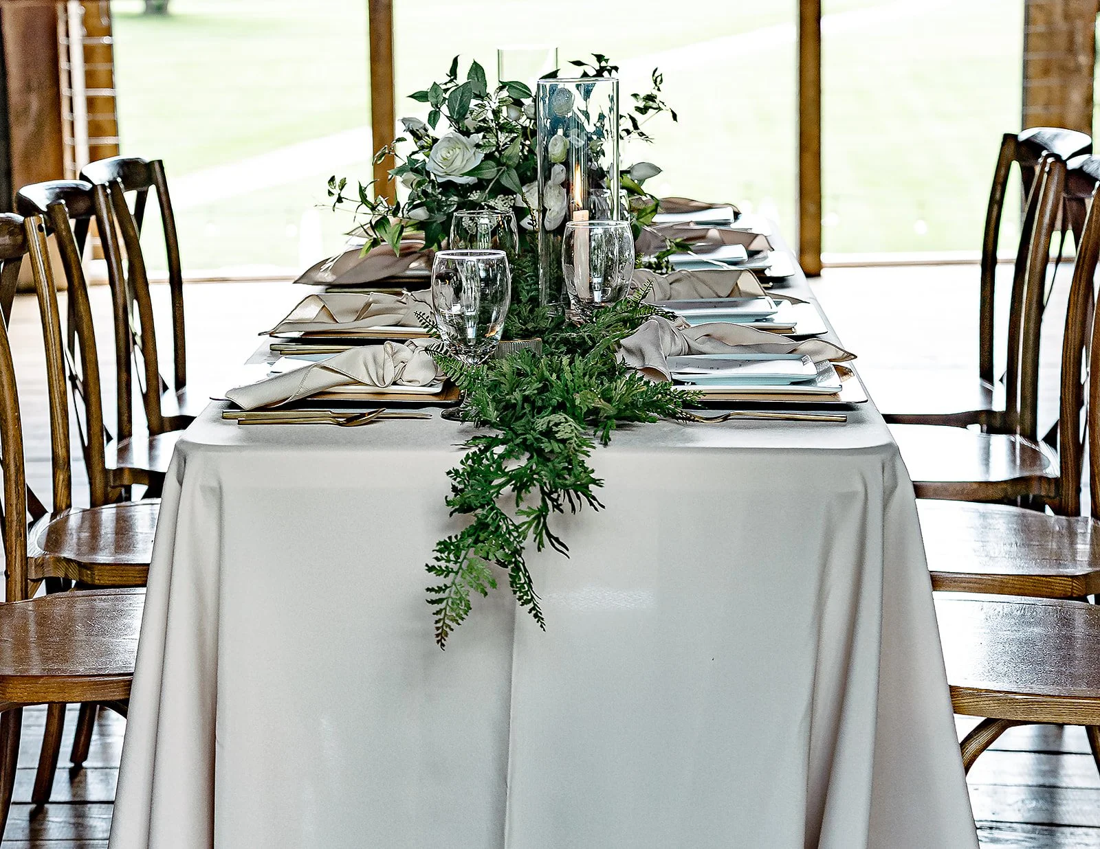 A beautifully decorated banquet table with a white tablecloth, centerpiece with white flowers and greenery, surrounded by wooden chairs, in a bright room with large windows at a 19th century estate
