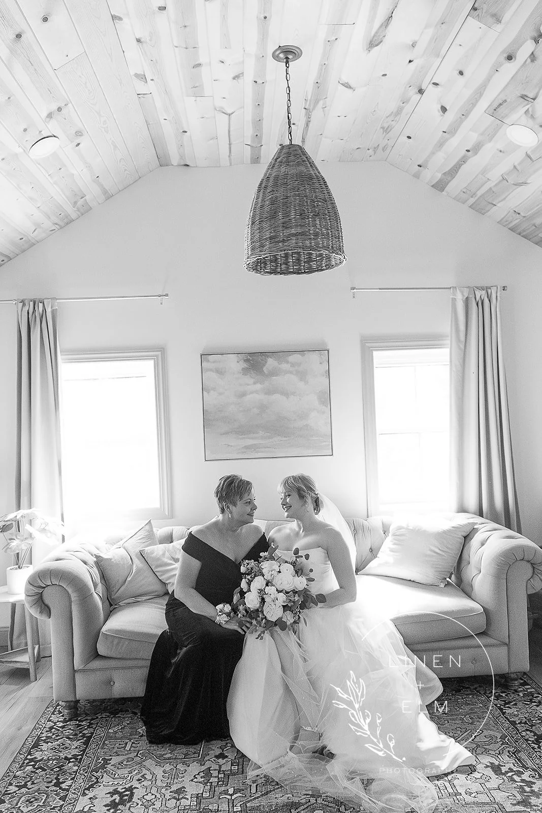 Black and white photo of two women sitting on a couch, one in a wedding dress holding a bouquet, the other in a black dress, smiling at each other, in a room with wooden ceiling and two windows.