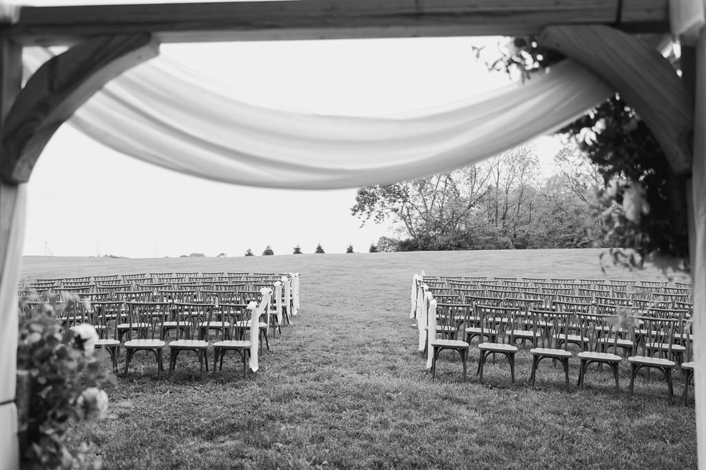 Empty chairs arranged for an outdoor wedding ceremony under a decorated canopy on a grassy field with trees in the background at a 19th century estate