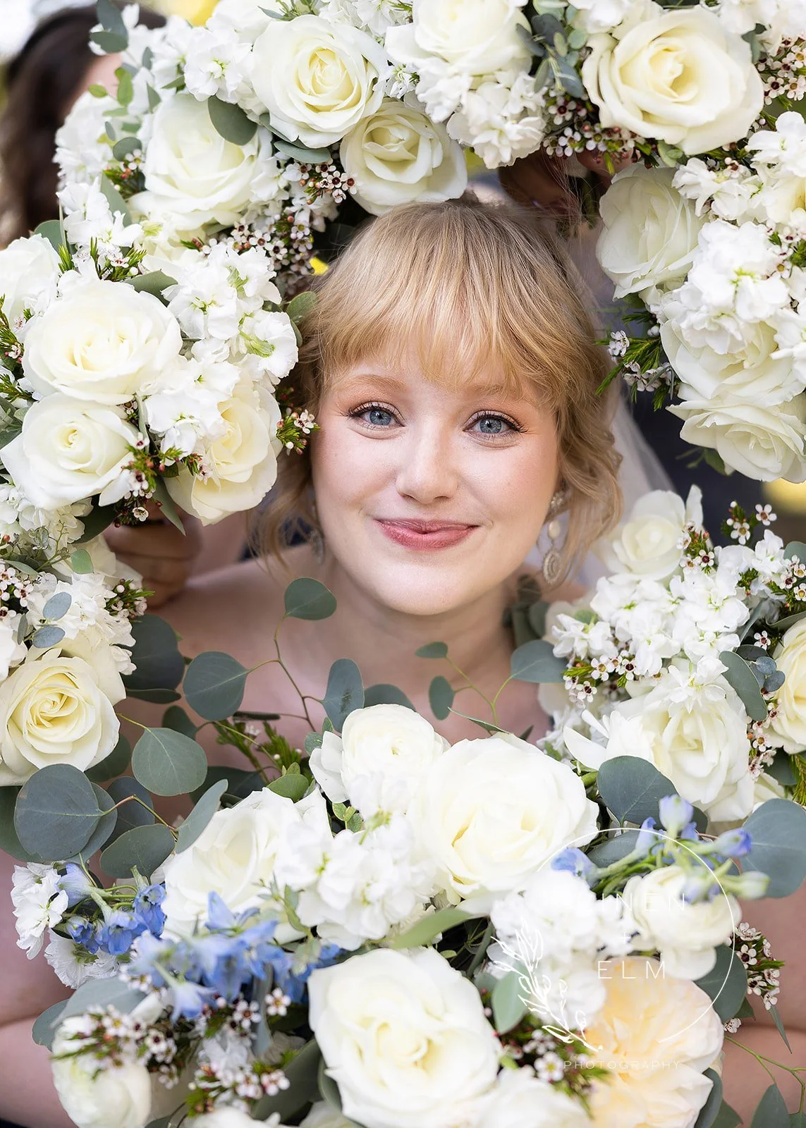 A woman with blonde hair and blue eyes smiling, surrounded by a large bouquet of white roses, greenery, and small white and blue flowers.