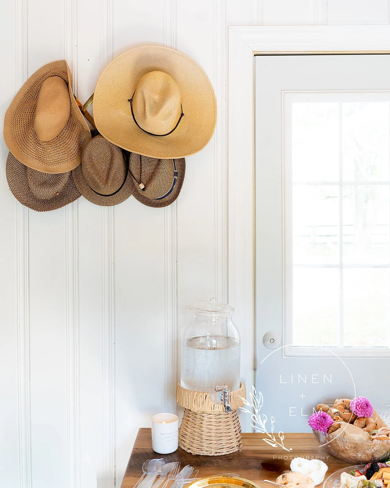 Six wide-brimmed straw hats hanging on a white paneled wall near a door with glass panes, with a water dispenser and a candle on a wooden table below.