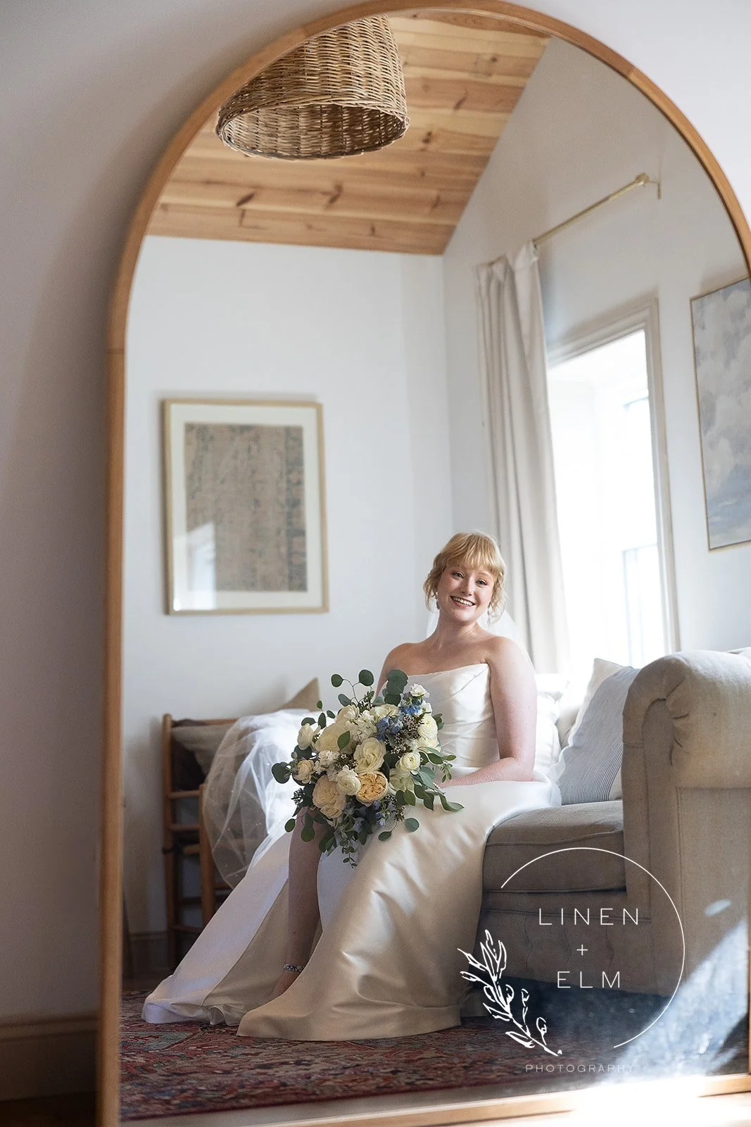 A woman in a wedding dress smiling while holding a bouquet, sitting on a sofa in a cozy, well-lit room, reflected in a full-length mirror with a wooden frame.