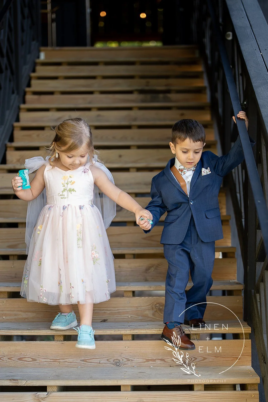 Two children, a girl in a white dress with floral embroidery and a boy in a blue suit, are holding hands and descending wooden stairs at a formal event.