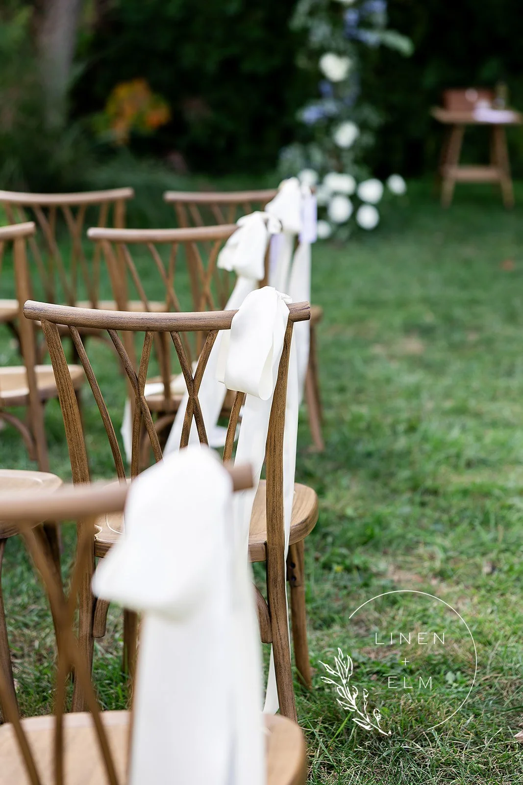 Wooden chairs decorated with white ribbons arranged in a row outdoors, likely for a wedding or special event with greenery in the background.